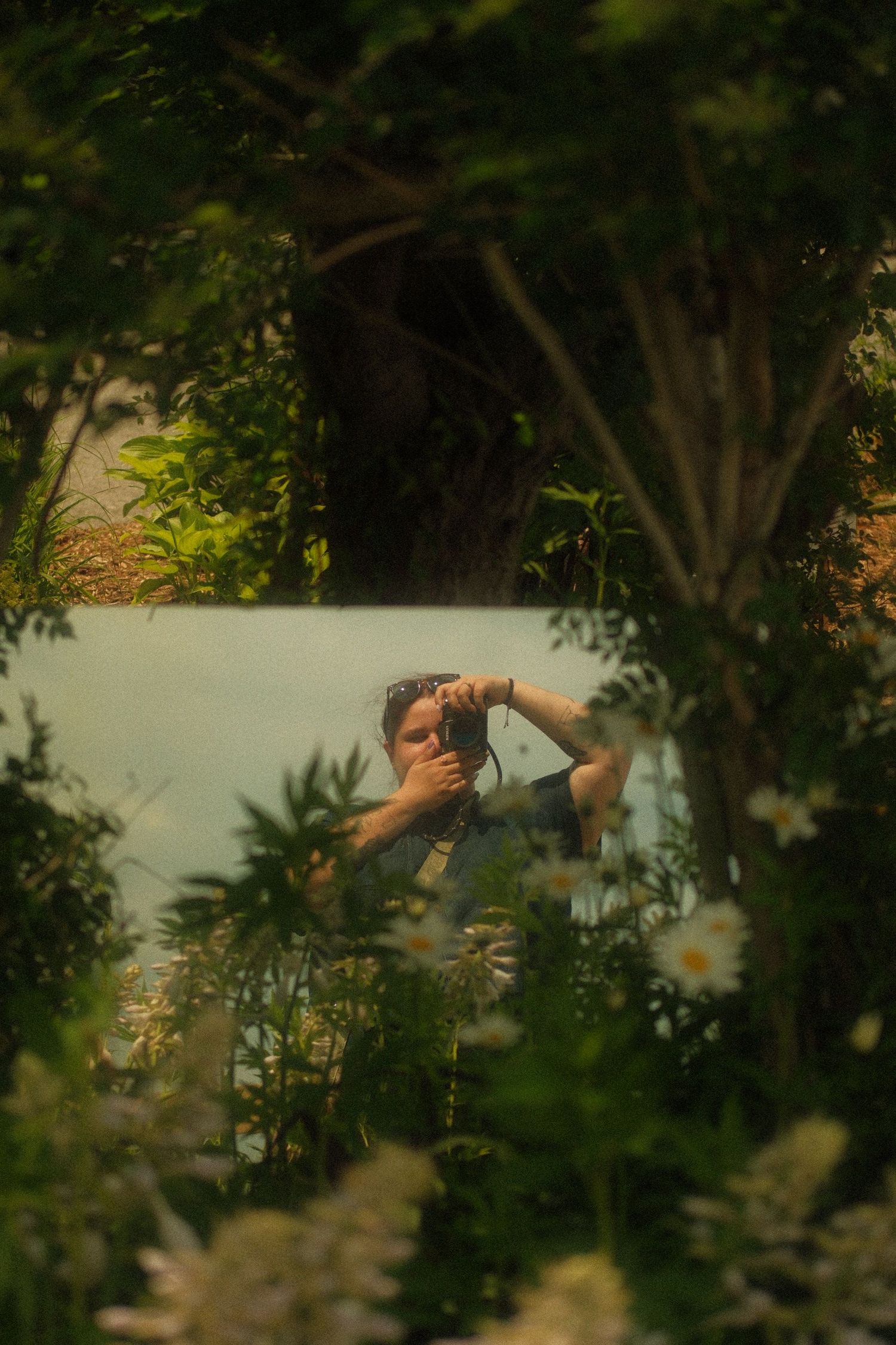 A photographer captures their reflection in a mirror surrounded by lush green garden foliage and white flowers.