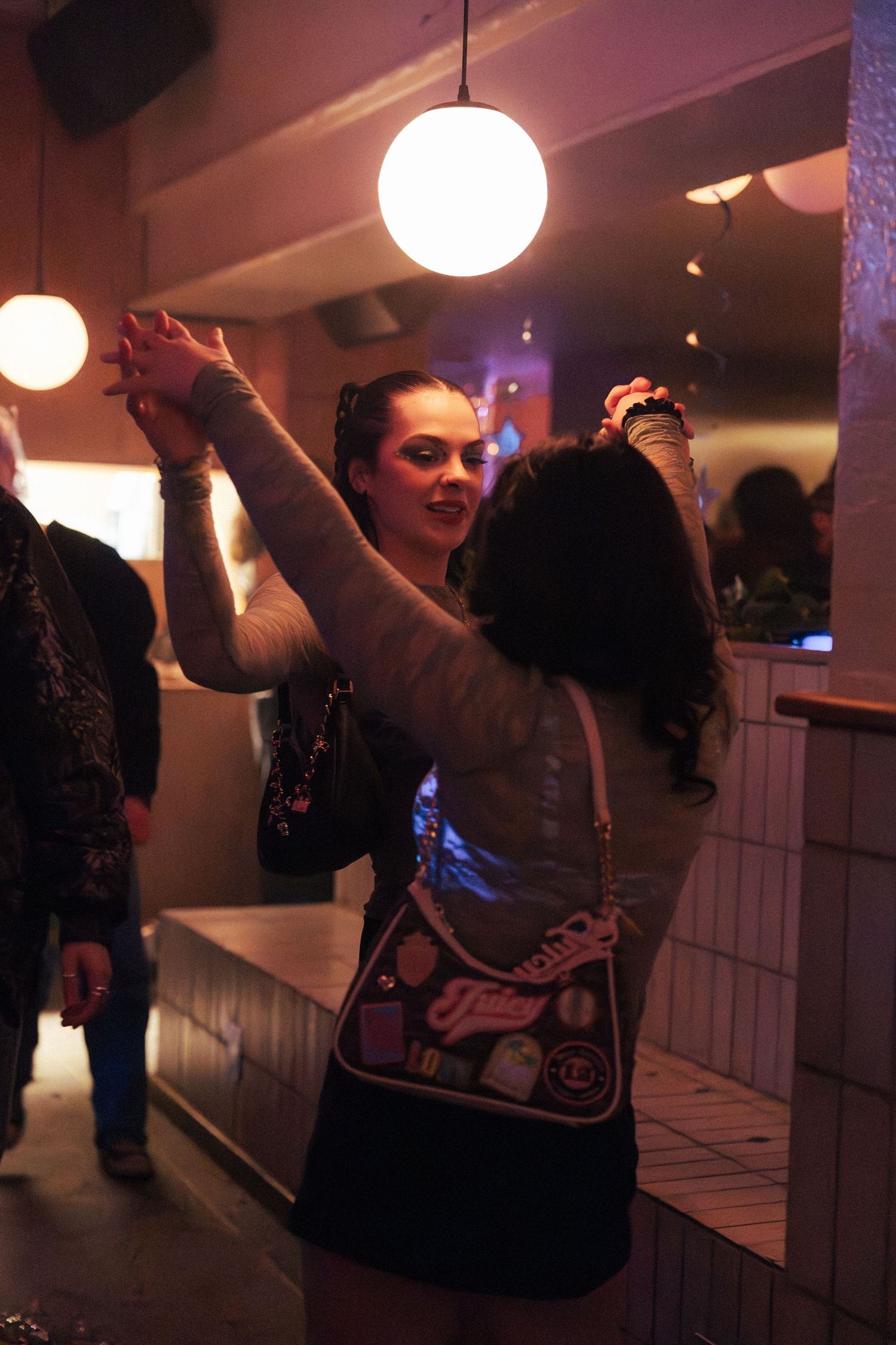 Two women dancing together at a nightclub under warm globe lights, one wearing a designer handbag.
