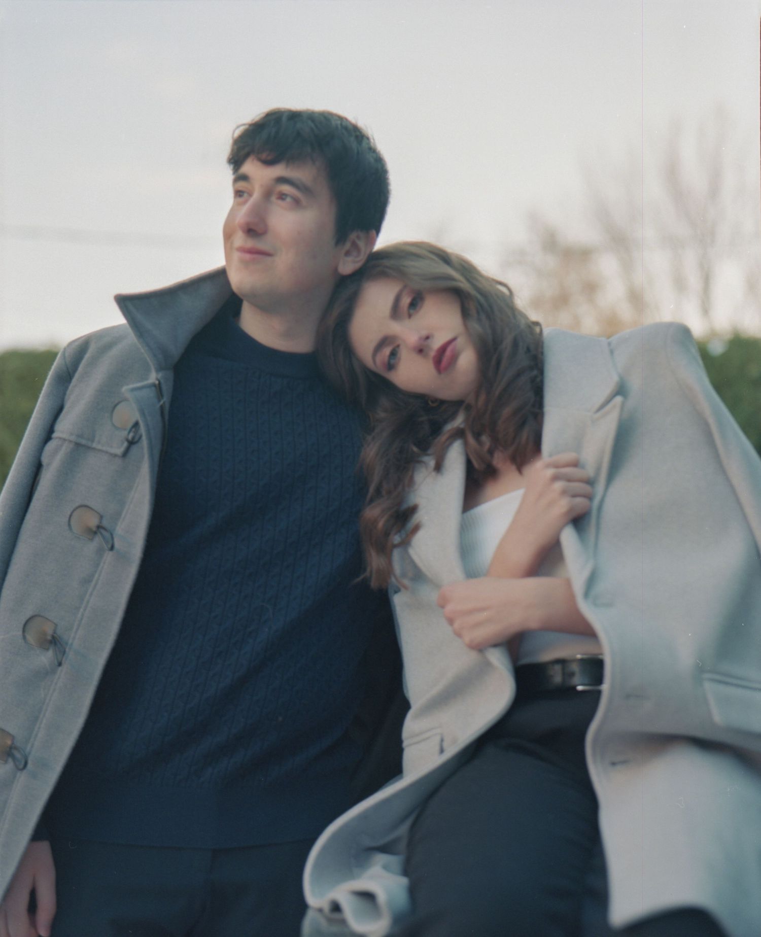 A young couple leans together outdoors in matching gray coats during golden hour.