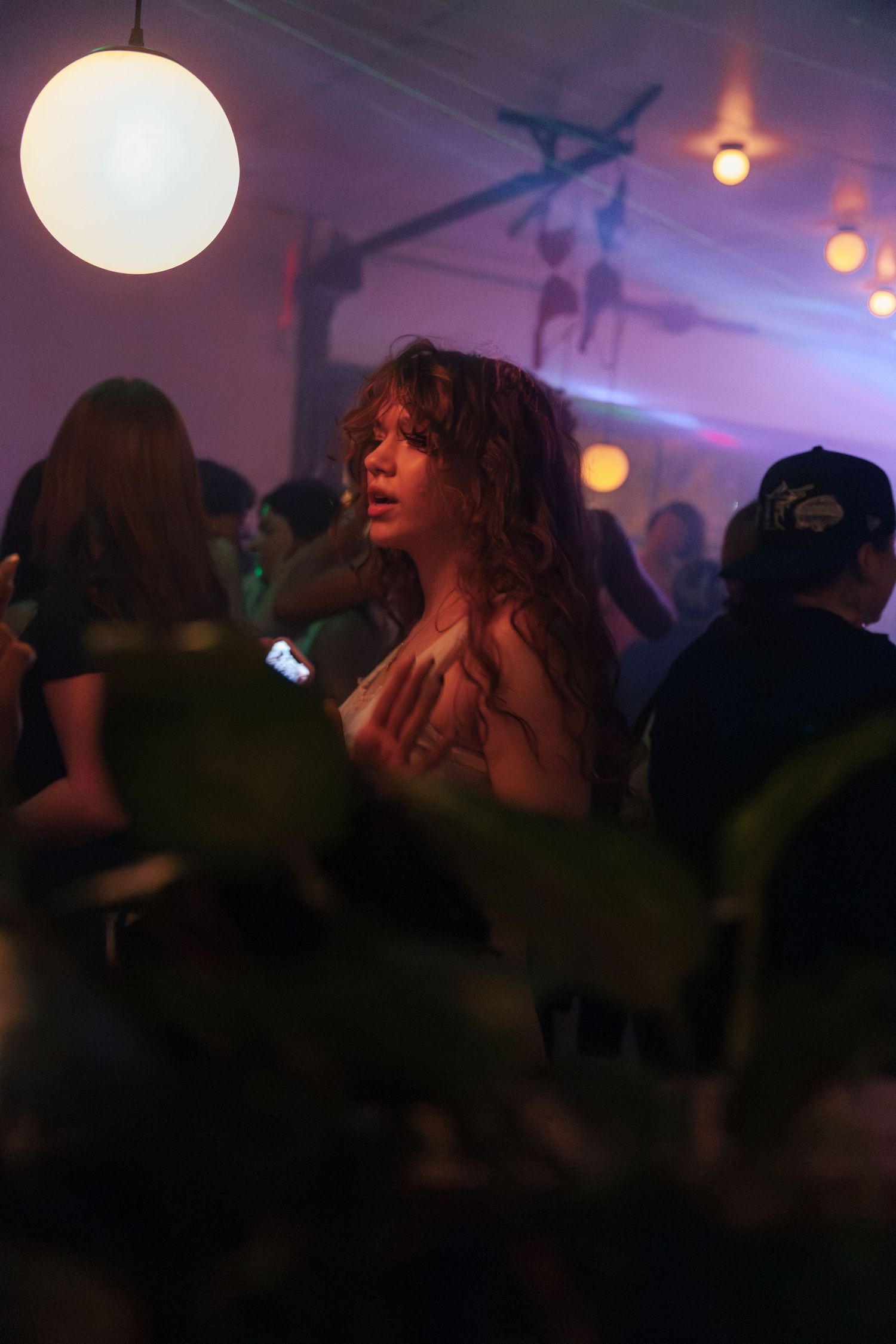 Woman with curly hair dancing at a dimly lit party with glowing lights and crowd in background.