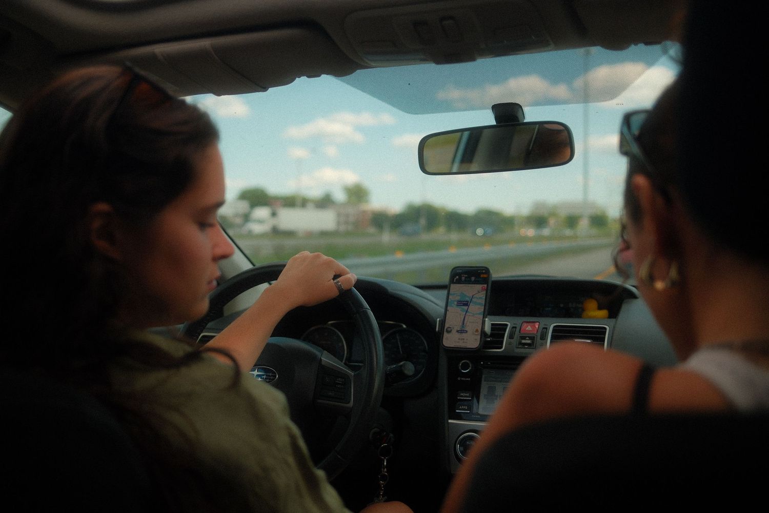 Two women inside a car, one driving while using a phone mounted on the dashboard for navigation.