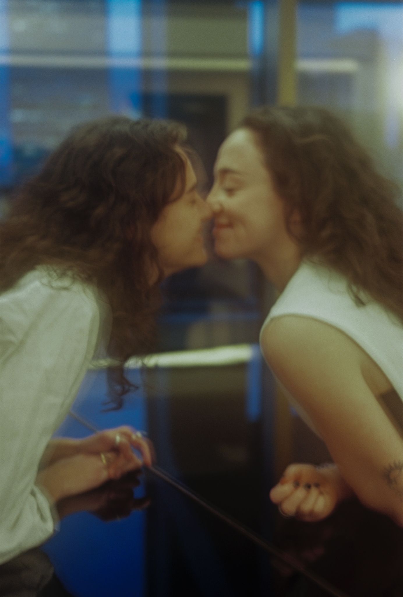 Two young women with curly hair share a romantic kiss in a dimly lit indoor setting.