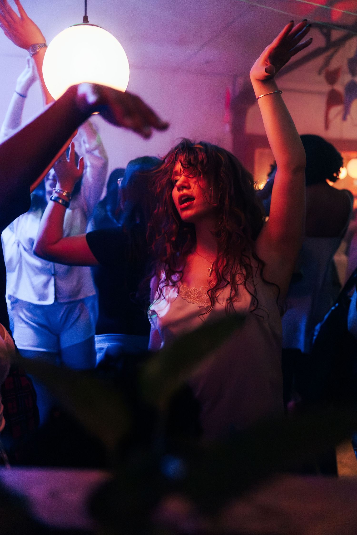 Young woman with curly hair dancing at a vibrant party with hands raised under glowing pendant light.