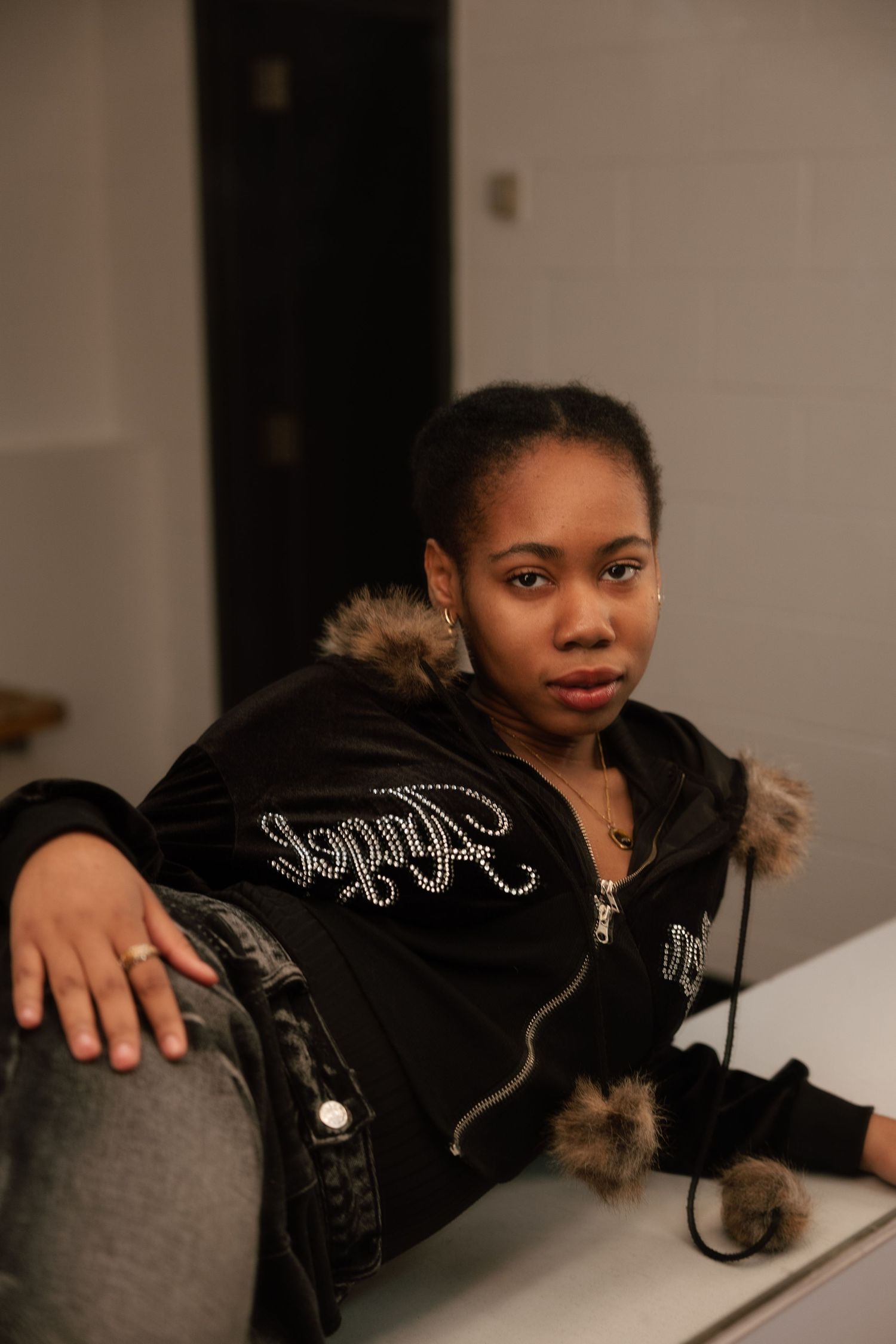 Young woman in black rhinestone jacket with fur trim leans on white surface, looking at camera.