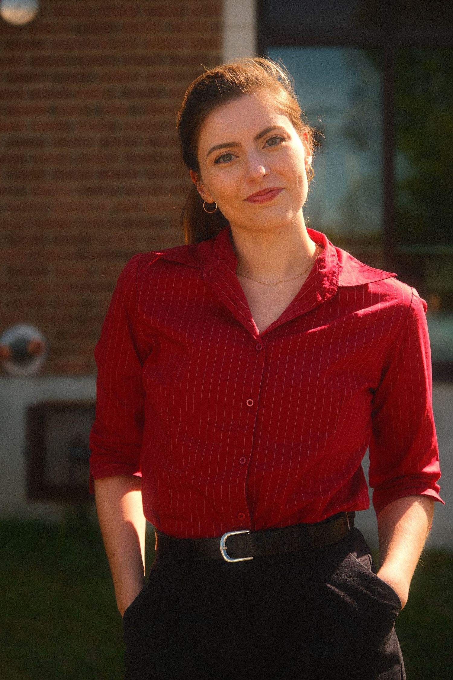 Young woman in red striped shirt and black pants standing outdoors near brick building in warm sunlight.