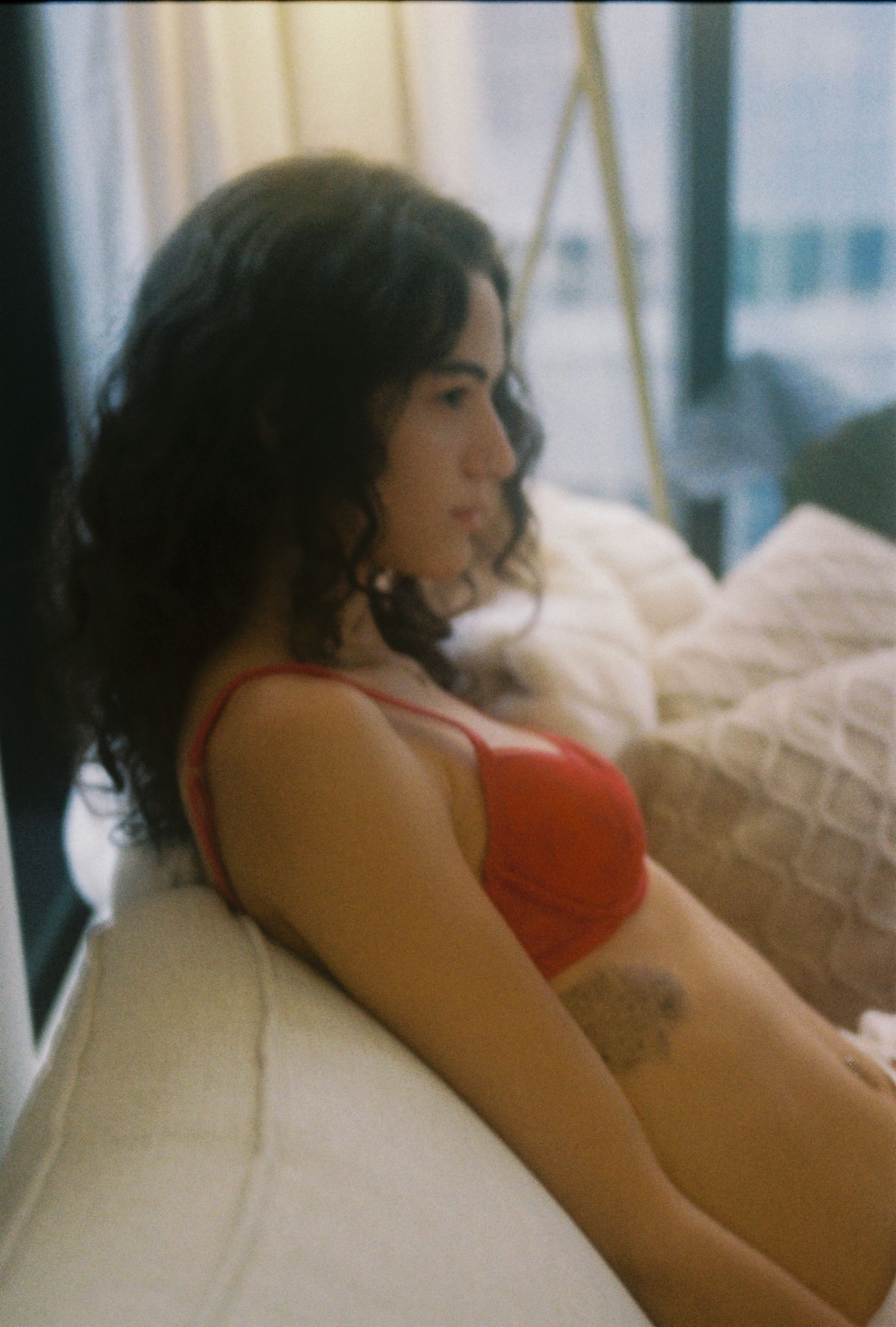 Woman in red bra sitting on bed with curly dark hair and tattoo.