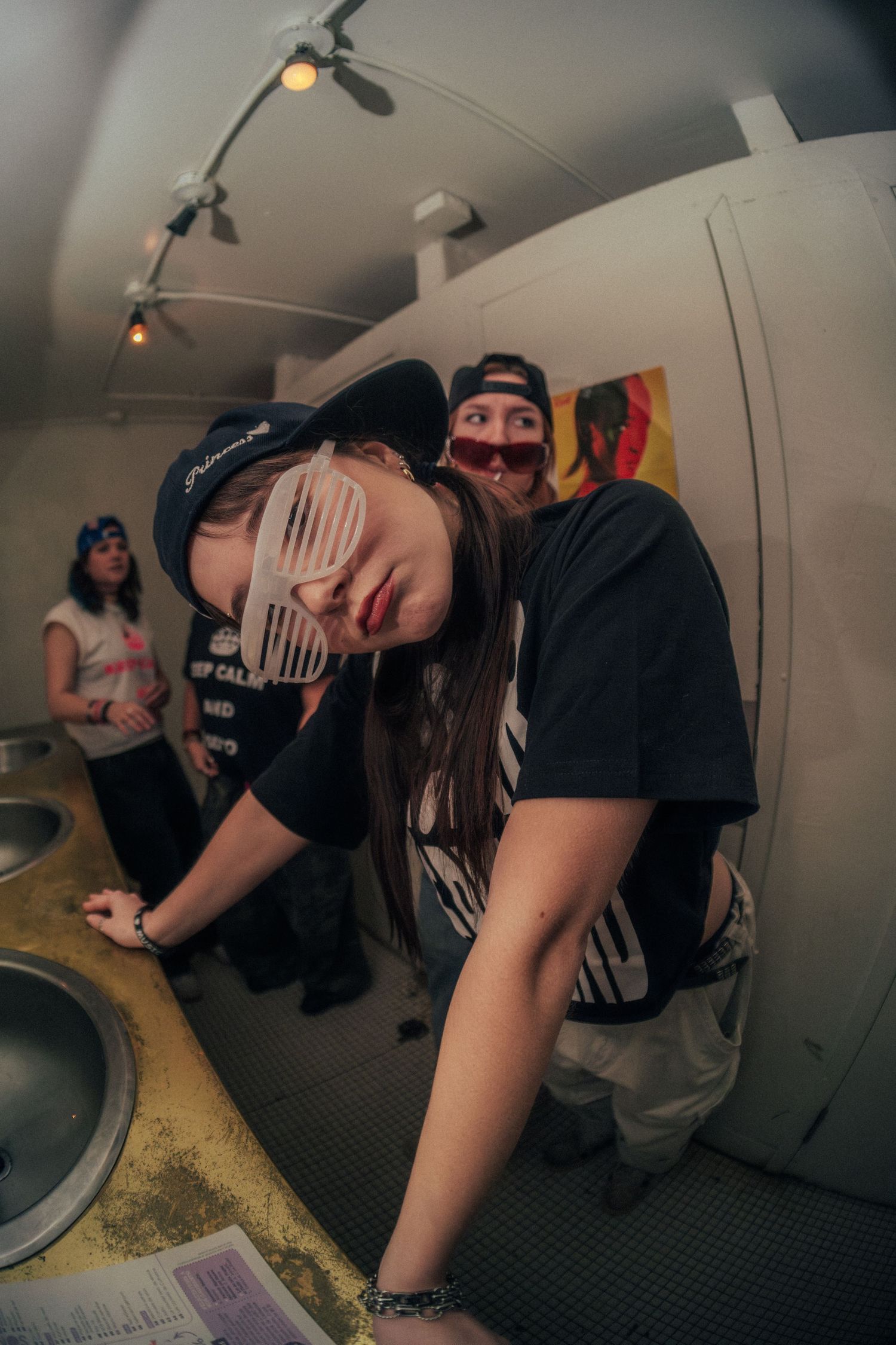 Person wearing shutter shades and snapback cap leaning over counter in dimly lit room with friends.