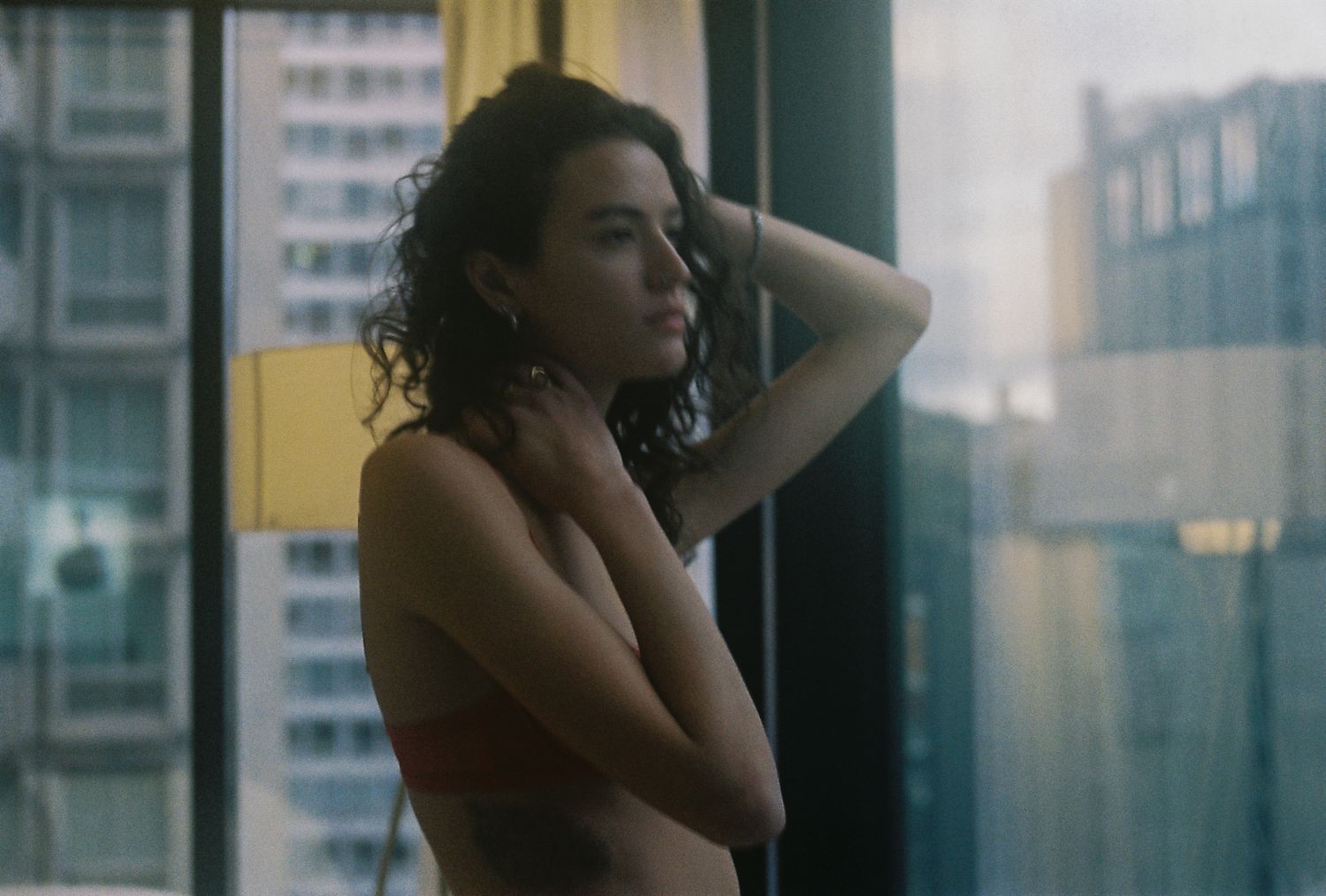A woman with curly hair stands by a window with a city skyline view, arms crossed in soft natural light.