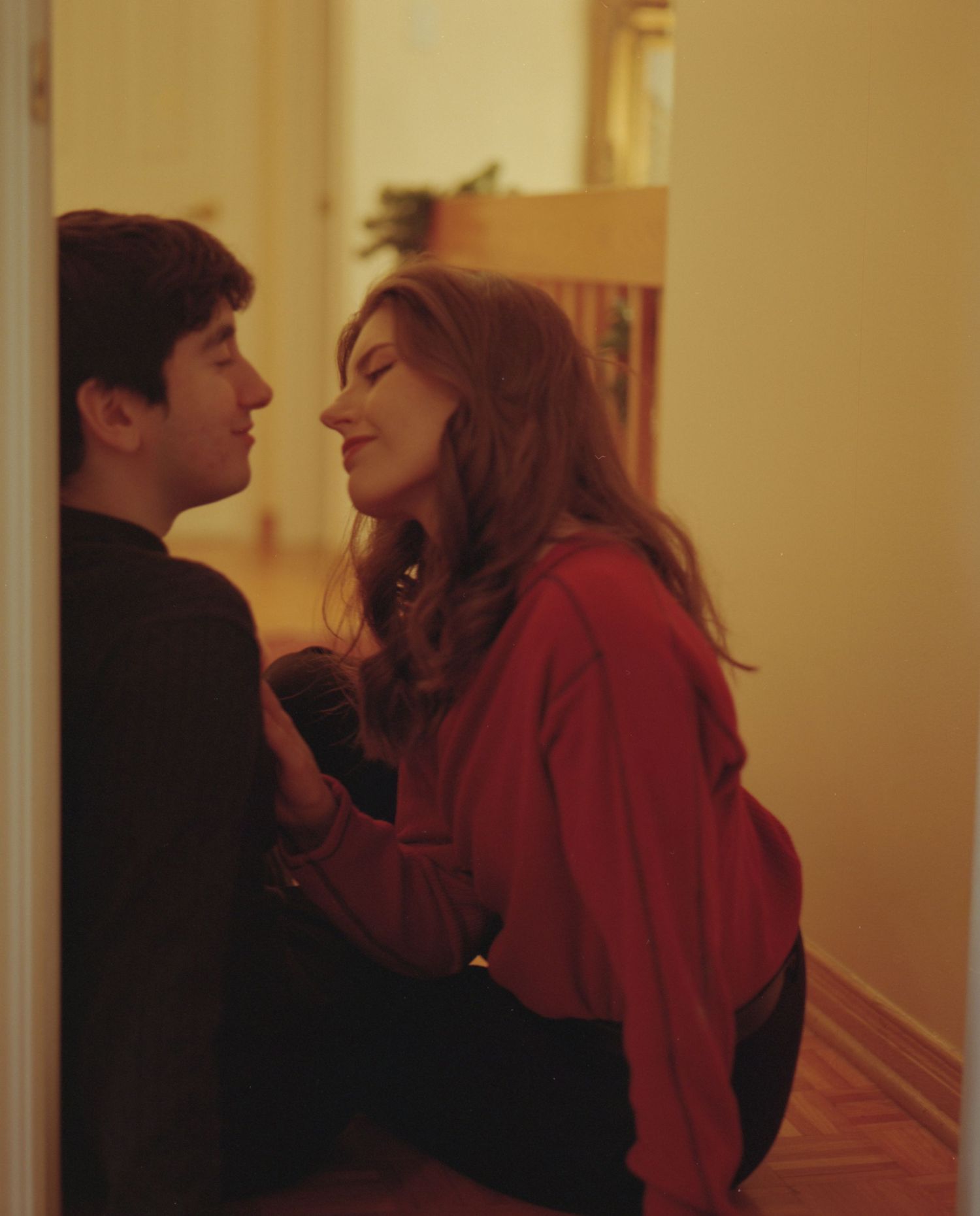A young couple shares an intimate moment leaning against a doorway in a warmly lit apartment.