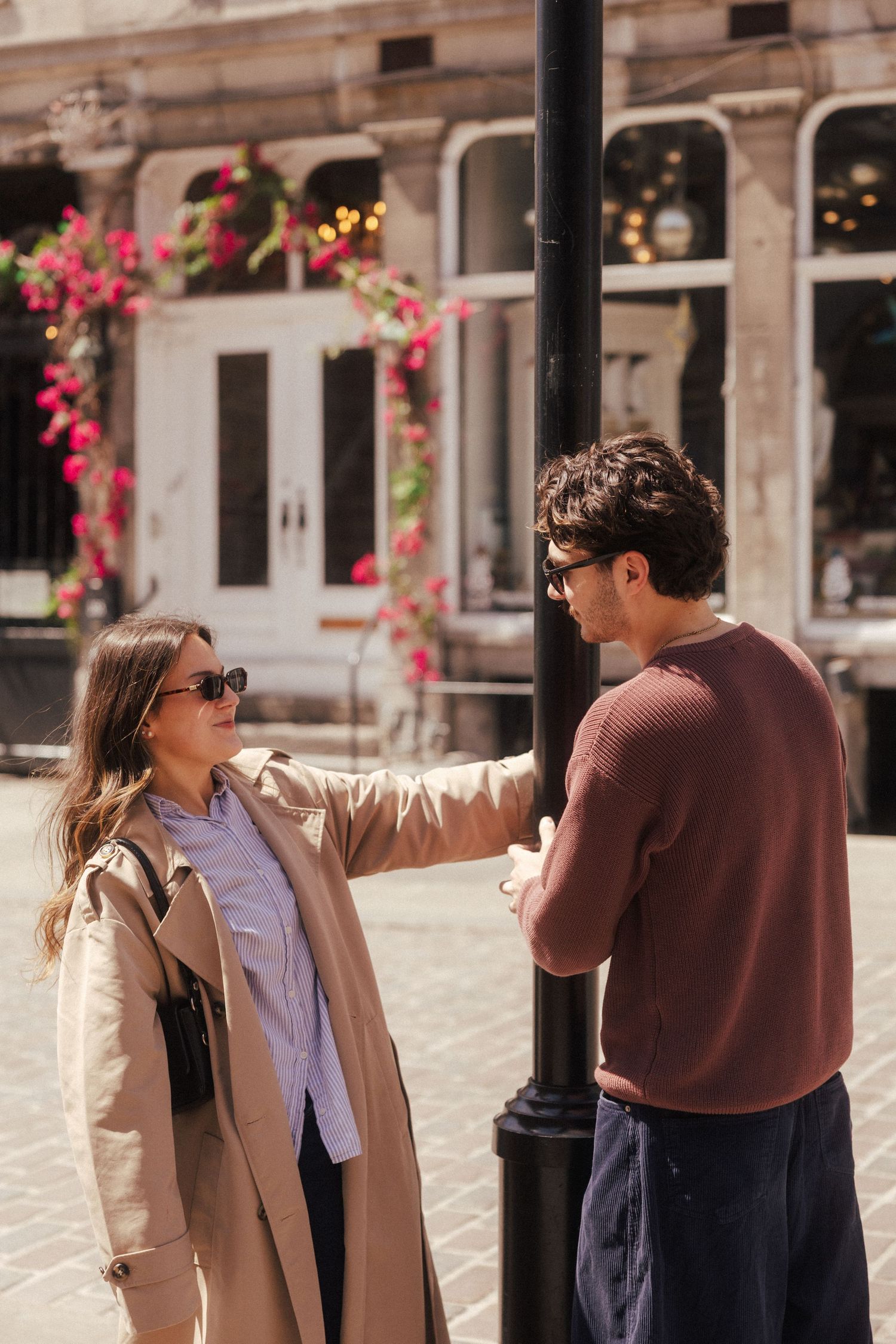 A man in a burgundy sweater gently touches a woman's shoulder on a sunny city street.