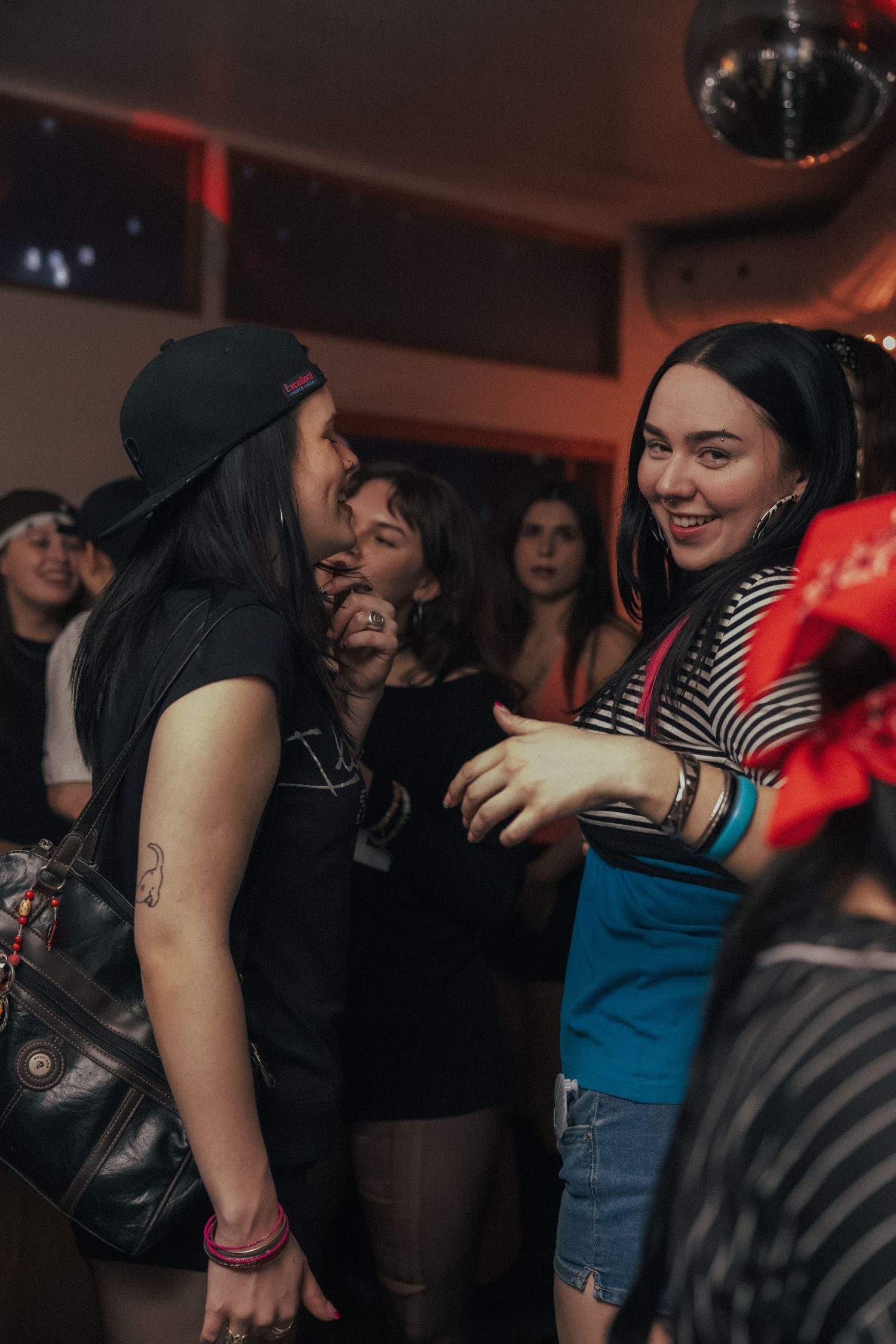 Two women smile and dance together at a lively nightclub with a disco ball overhead.
