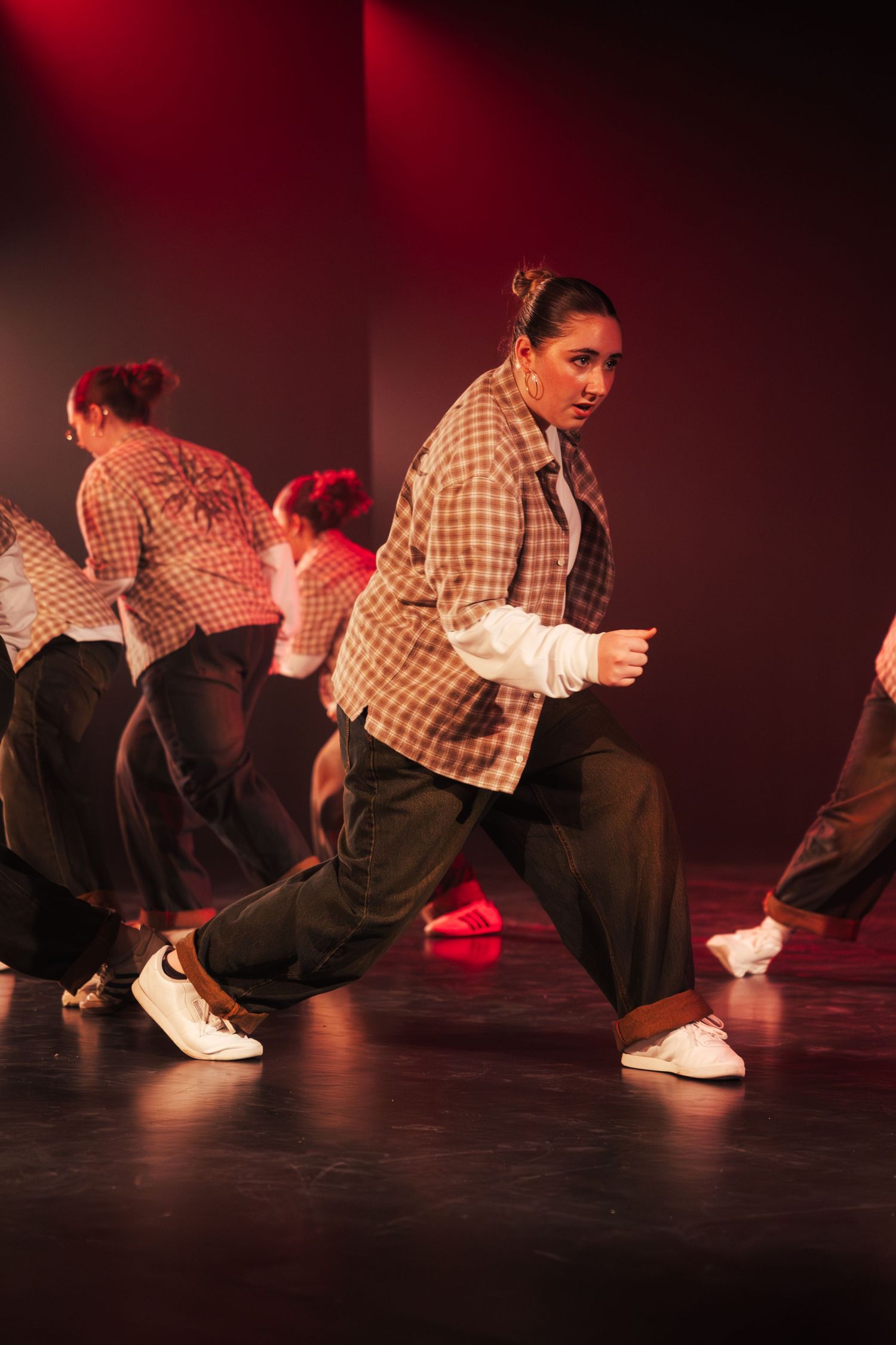 Dancers in matching plaid jackets and dark pants perform a hip-hop routine on a dimly lit stage with red lighting.