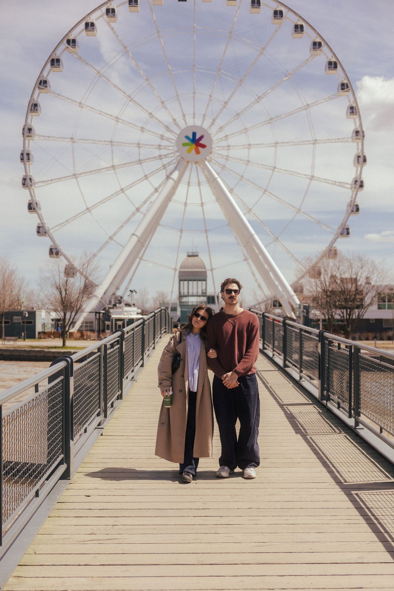 A couple poses together on a wooden bridge in front of a large Ferris wheel outdoors.