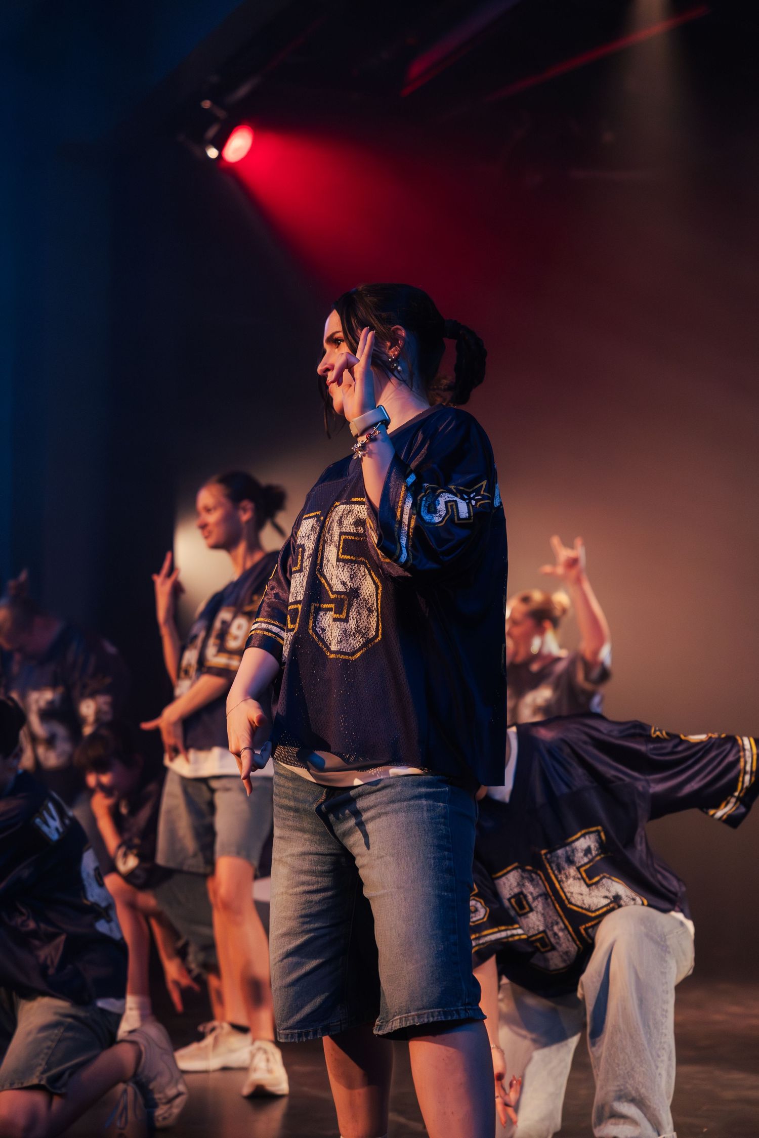 Smiling female dancer performing center stage in navy jersey number 95 with group dancers behind her under red lights.