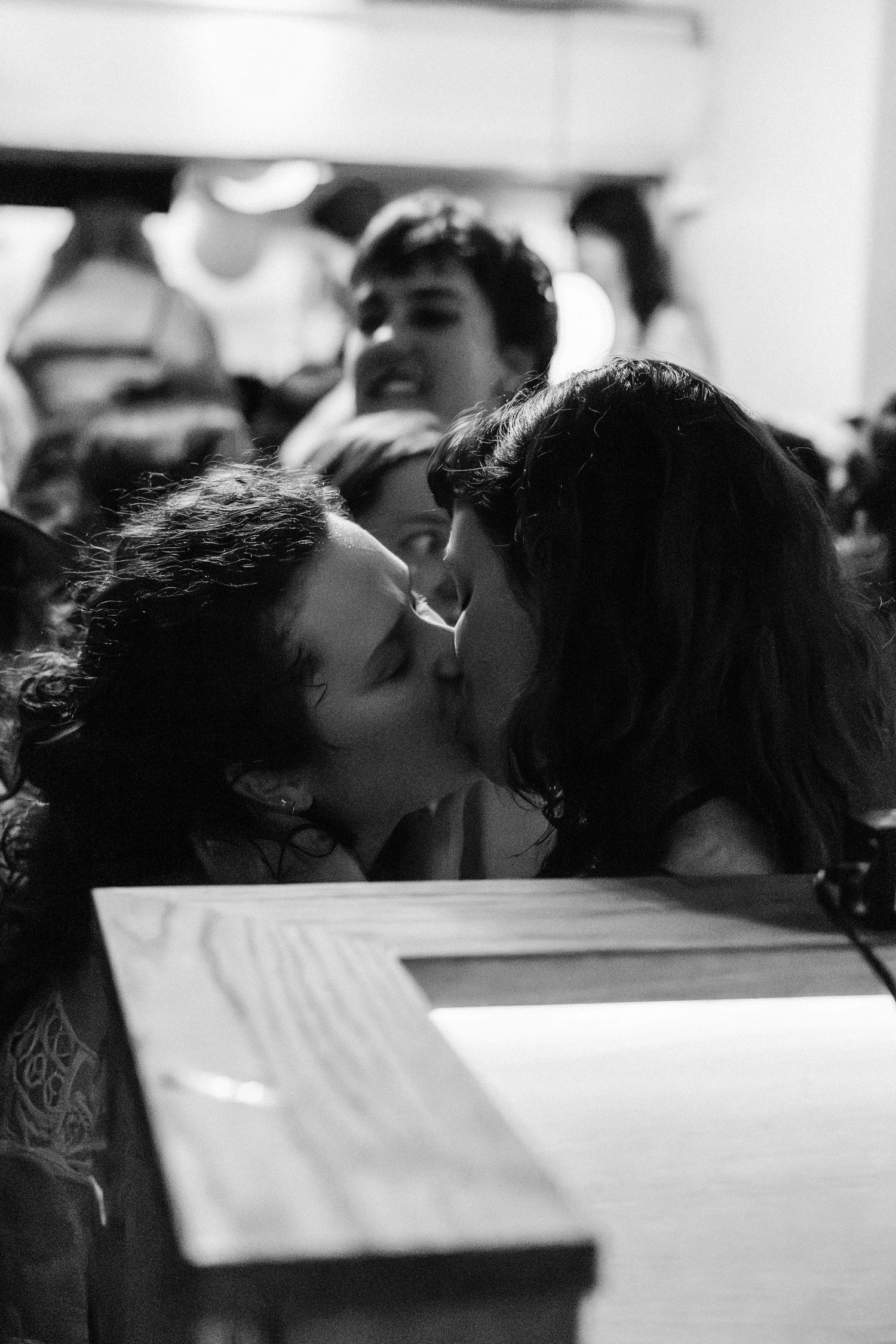 Black and white photo of two people sharing a kiss at a crowded indoor event.
