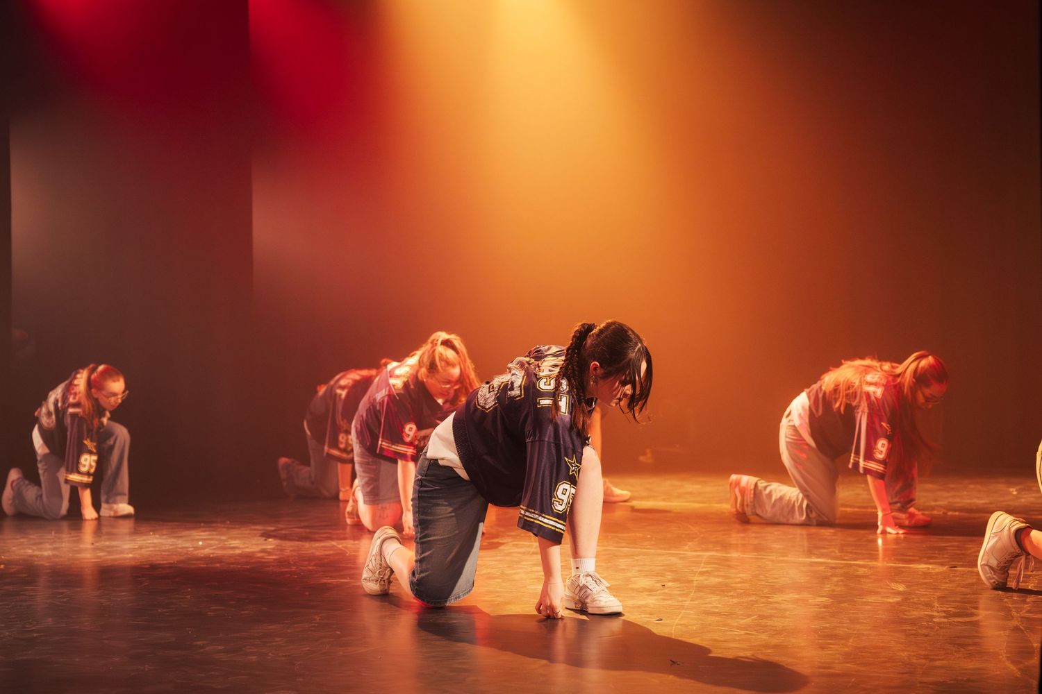 Hip-hop dancers kneeling on stage floor in casual streetwear during energetic group performance under warm lighting.