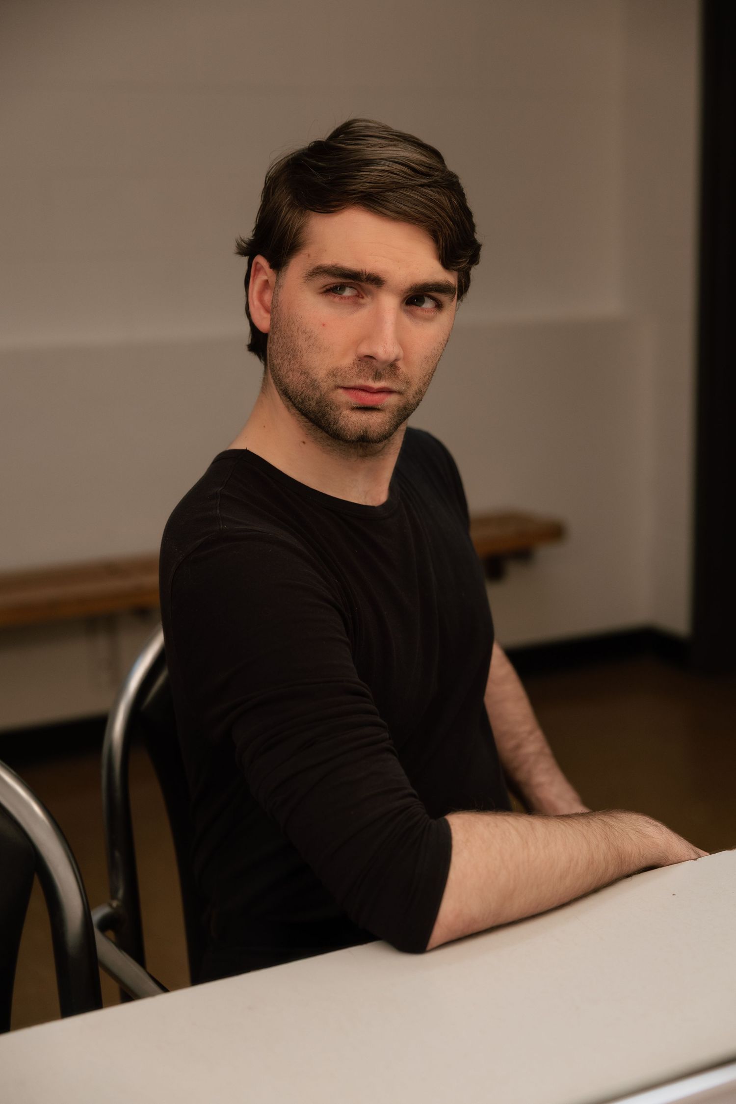 Young man with dark hair in black long-sleeve shirt sits at a white table, looking directly at camera.