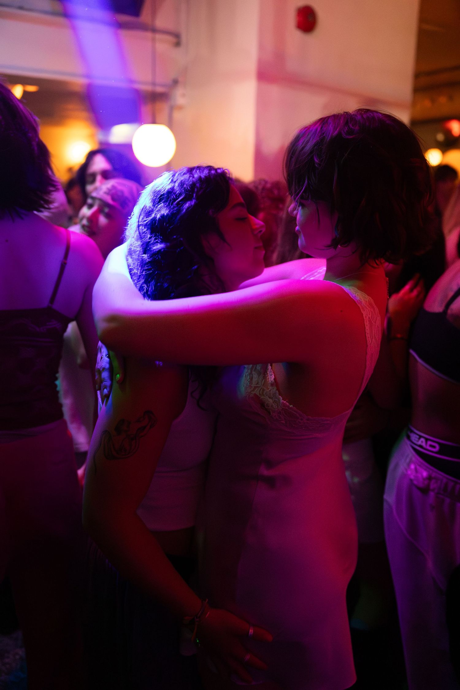 Two people slow dancing closely together under purple and pink lights on a crowded dance floor.