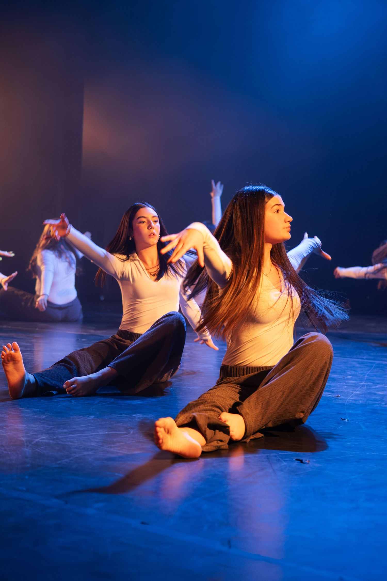 Female dancers sitting cross-legged on stage floor with arms extended during contemporary dance performance under blue lights.