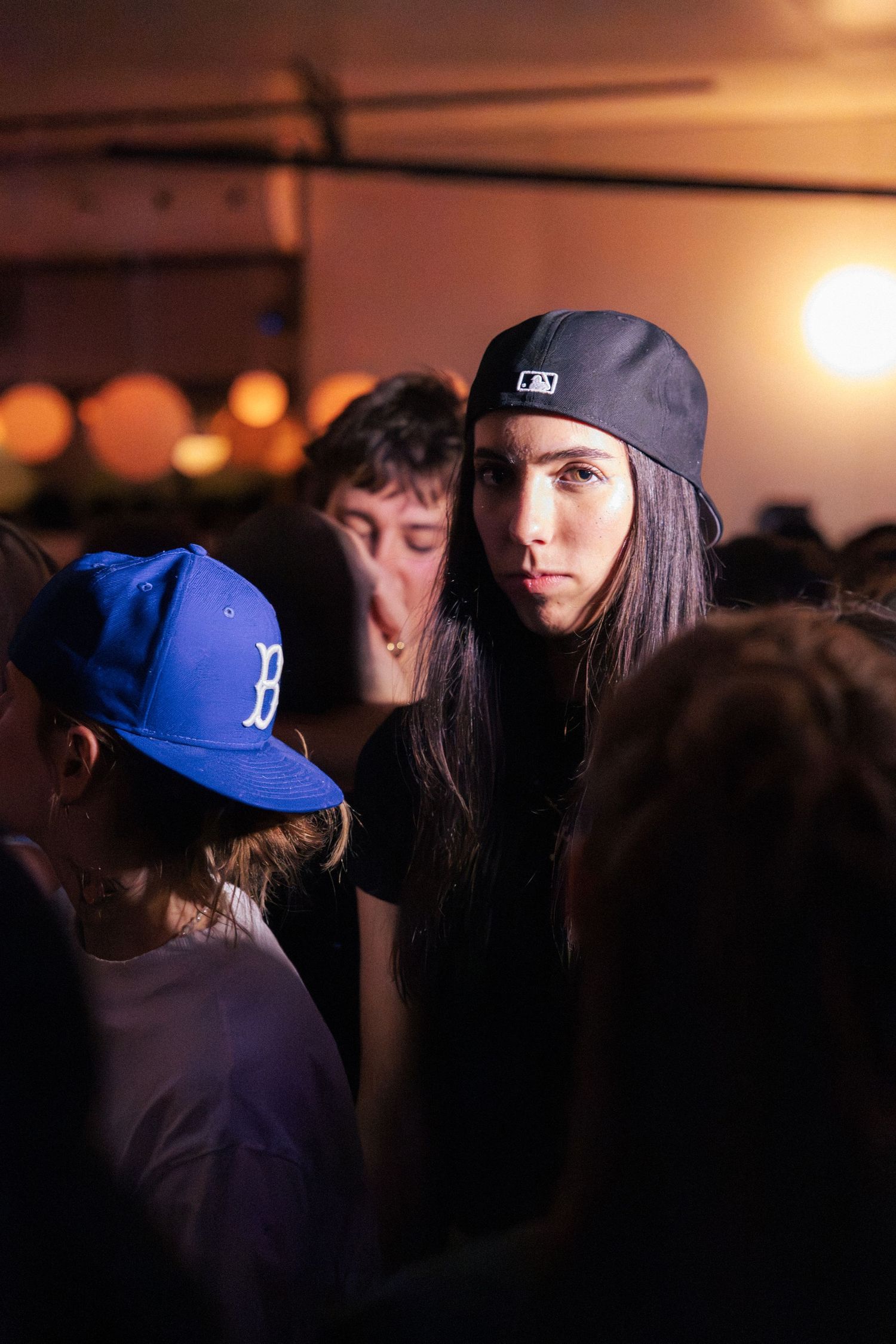 Two people wearing baseball caps stand in a warm-lit crowded party venue.