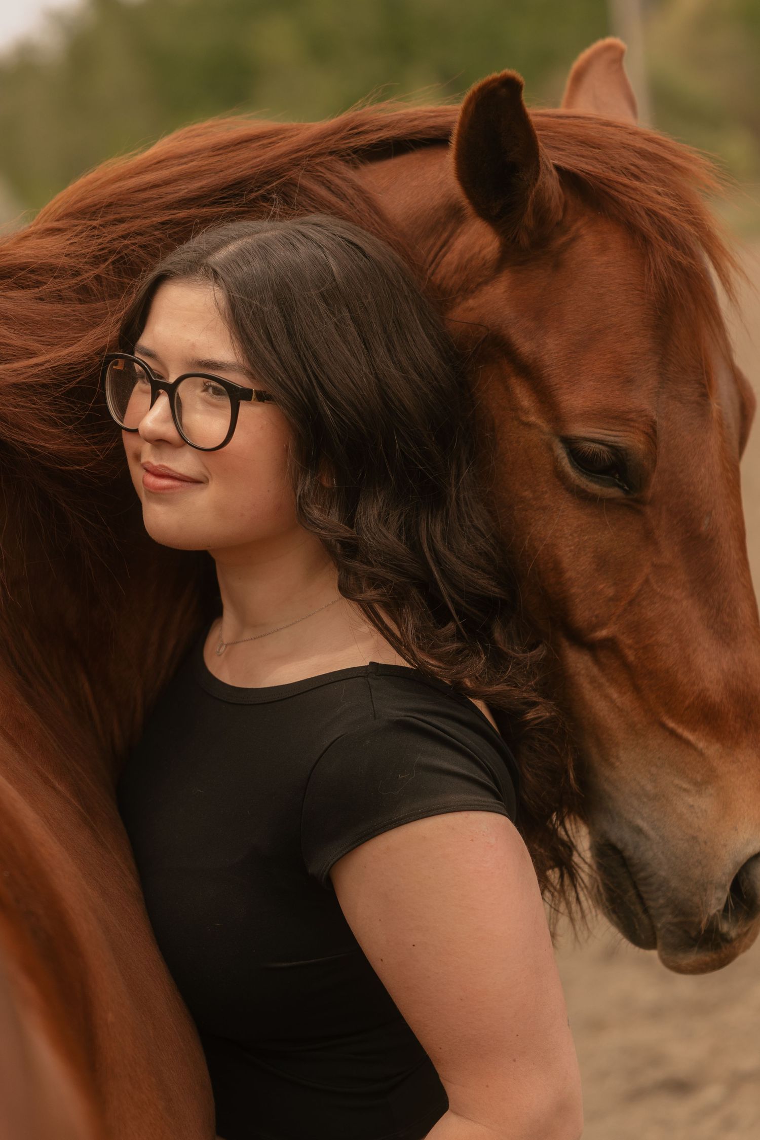 Young woman with glasses in black top stands beside a chestnut horse, its mane flowing gracefully around her.