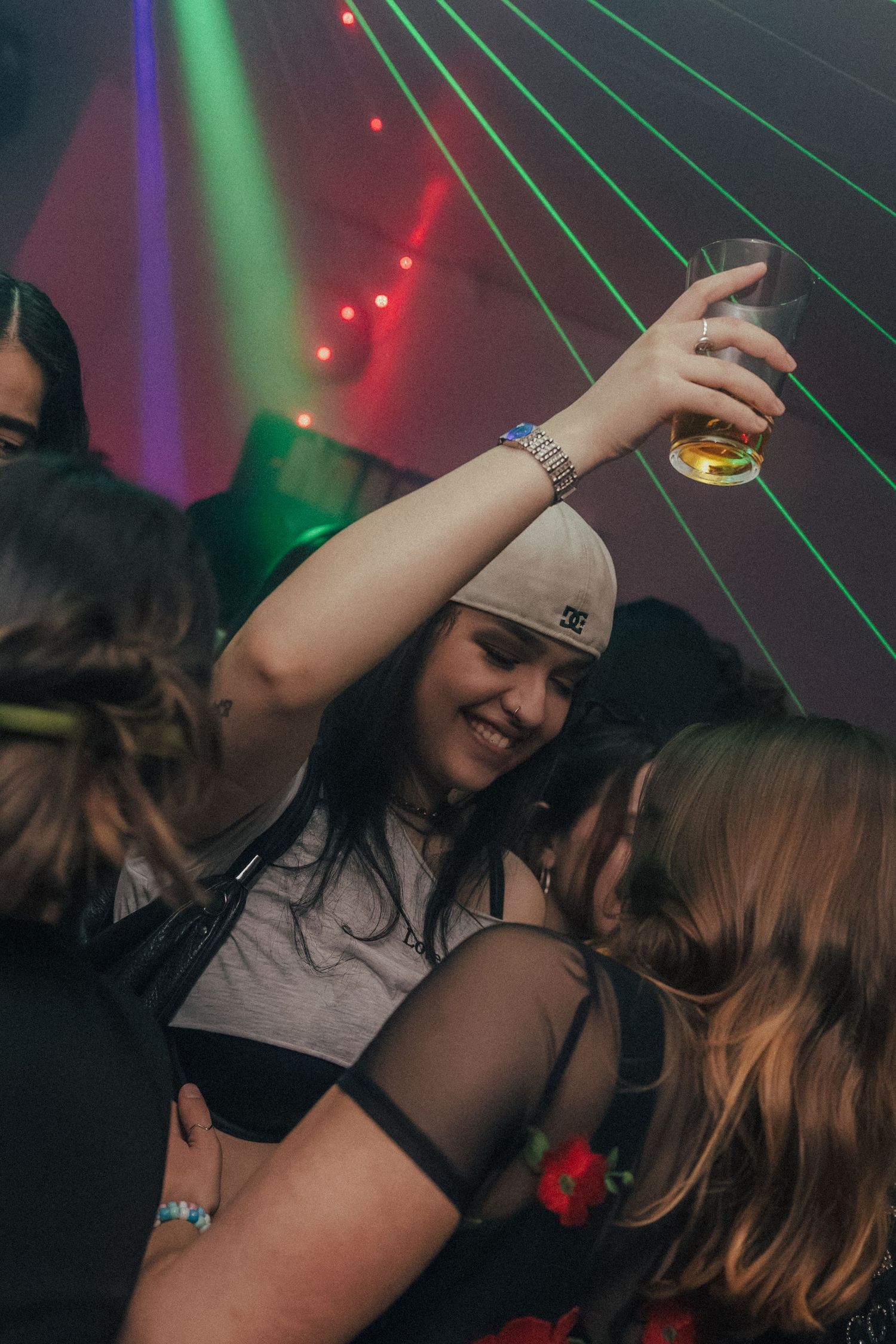 Woman in DC cap raises a drink while dancing under green laser lights at a nightclub.