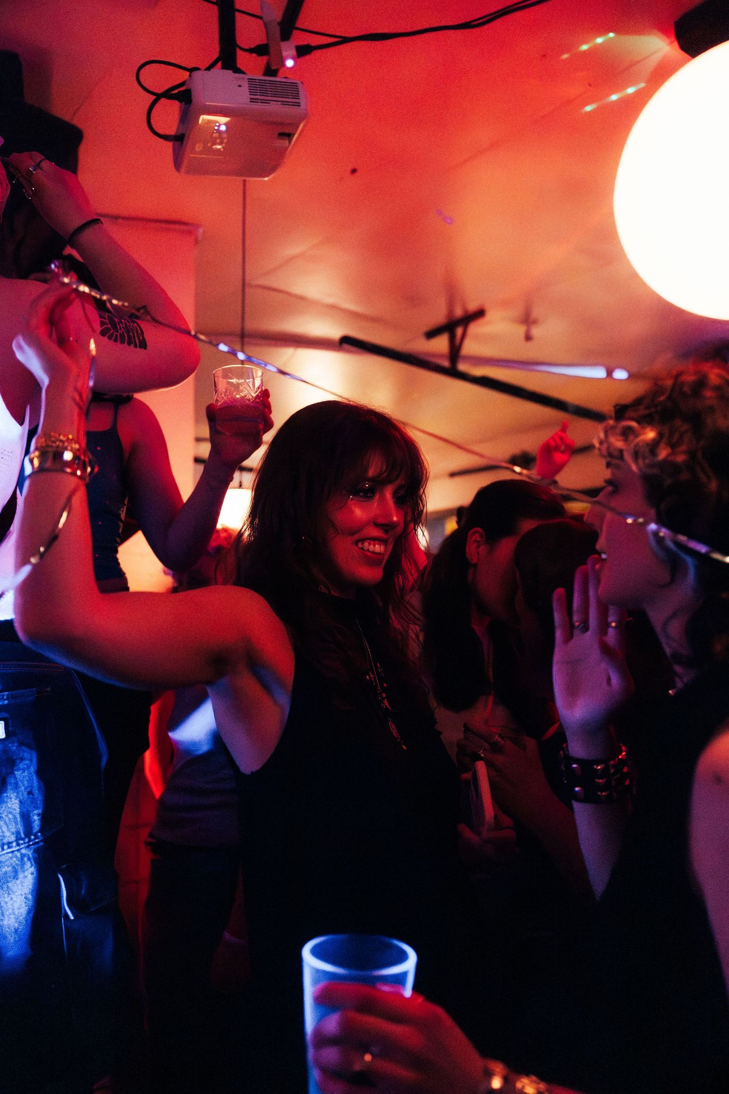 A smiling woman dances joyfully holding a drink at a crowded red-lit party venue.