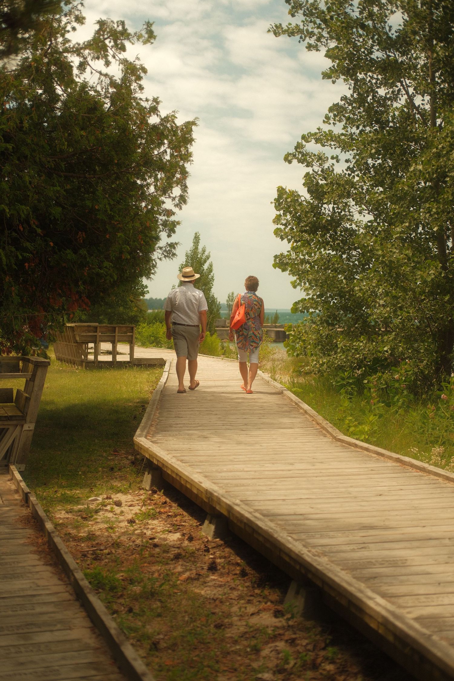 An elderly couple walks hand-in-hand along a wooden boardwalk path surrounded by lush green trees toward a lake.