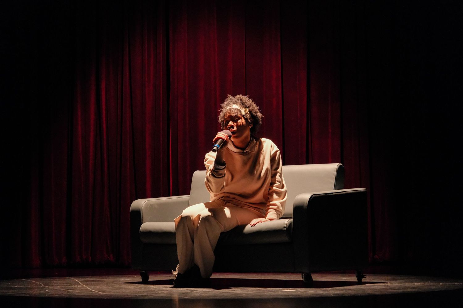 Performer sitting on white sofa holding microphone on stage with red curtain backdrop.