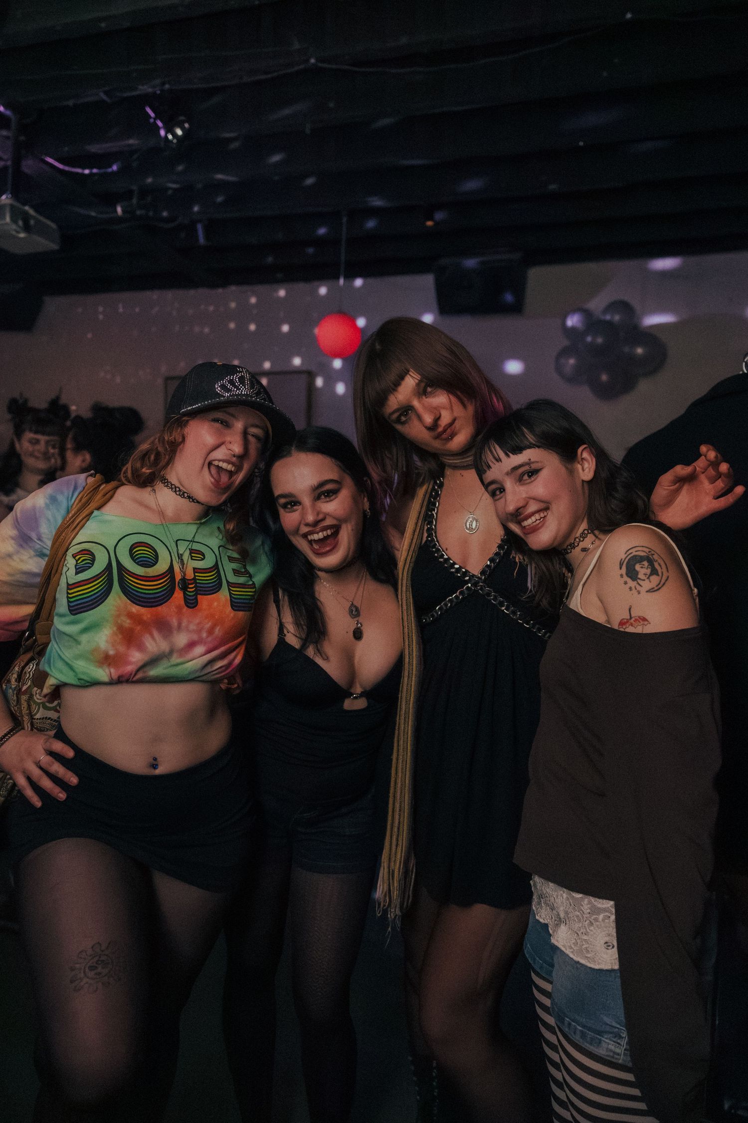 Four young women with tattoos smile and pose together on a nightclub dance floor.
