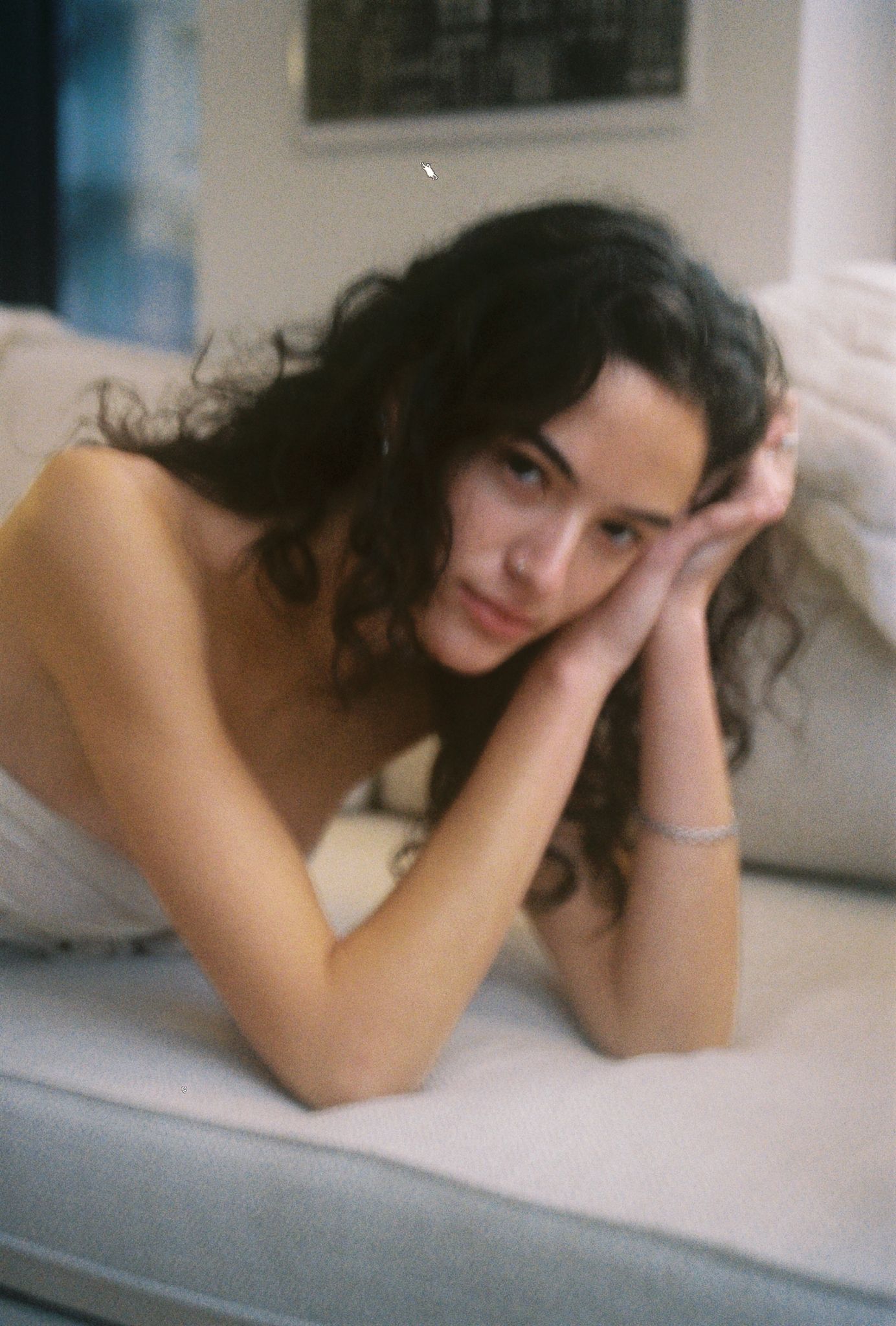 Young woman with curly dark hair resting her chin on her hands while lying on a white sofa indoors.