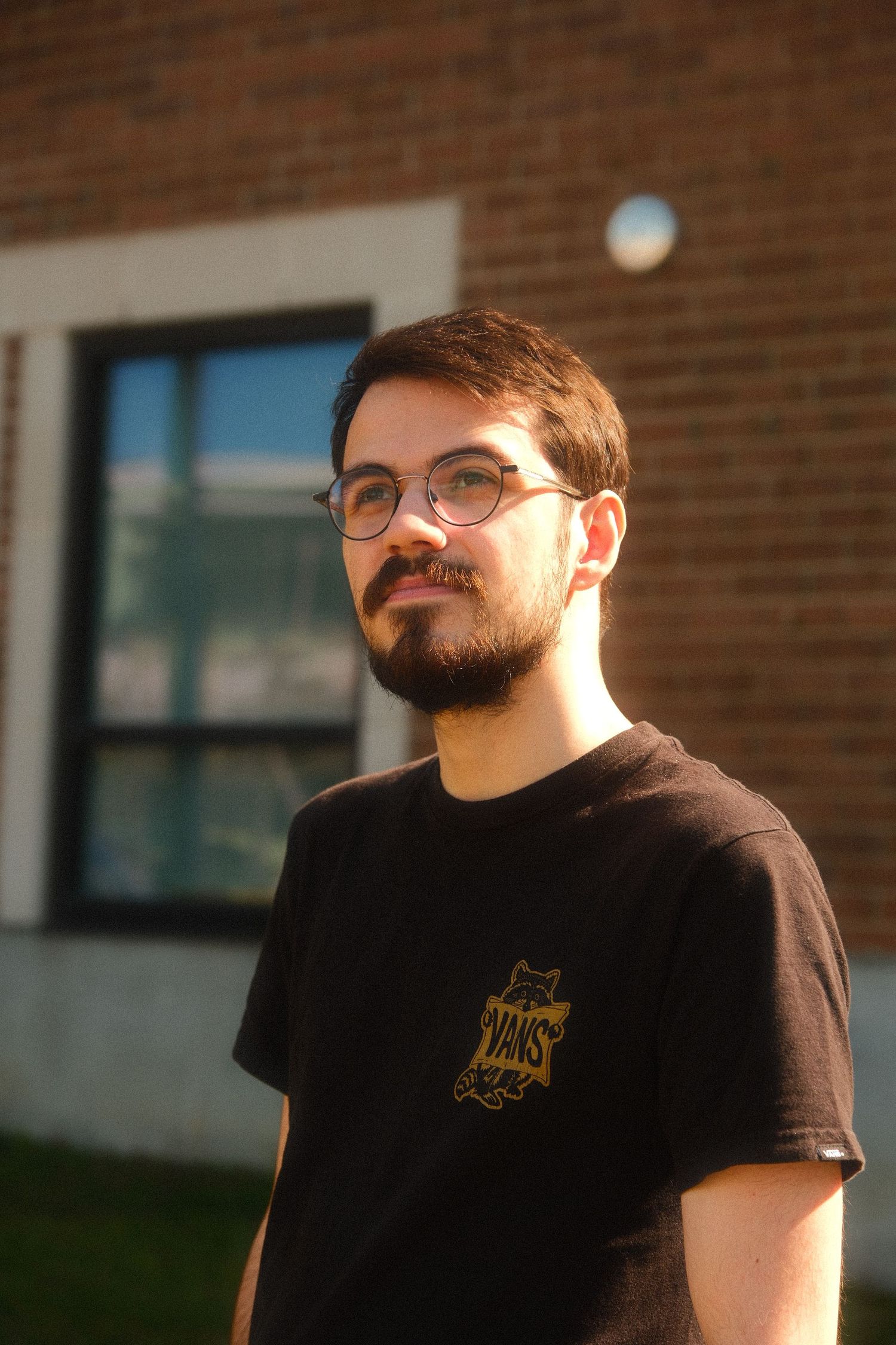 Young man with glasses and beard wearing a black Vans t-shirt standing outdoors near a brick building.