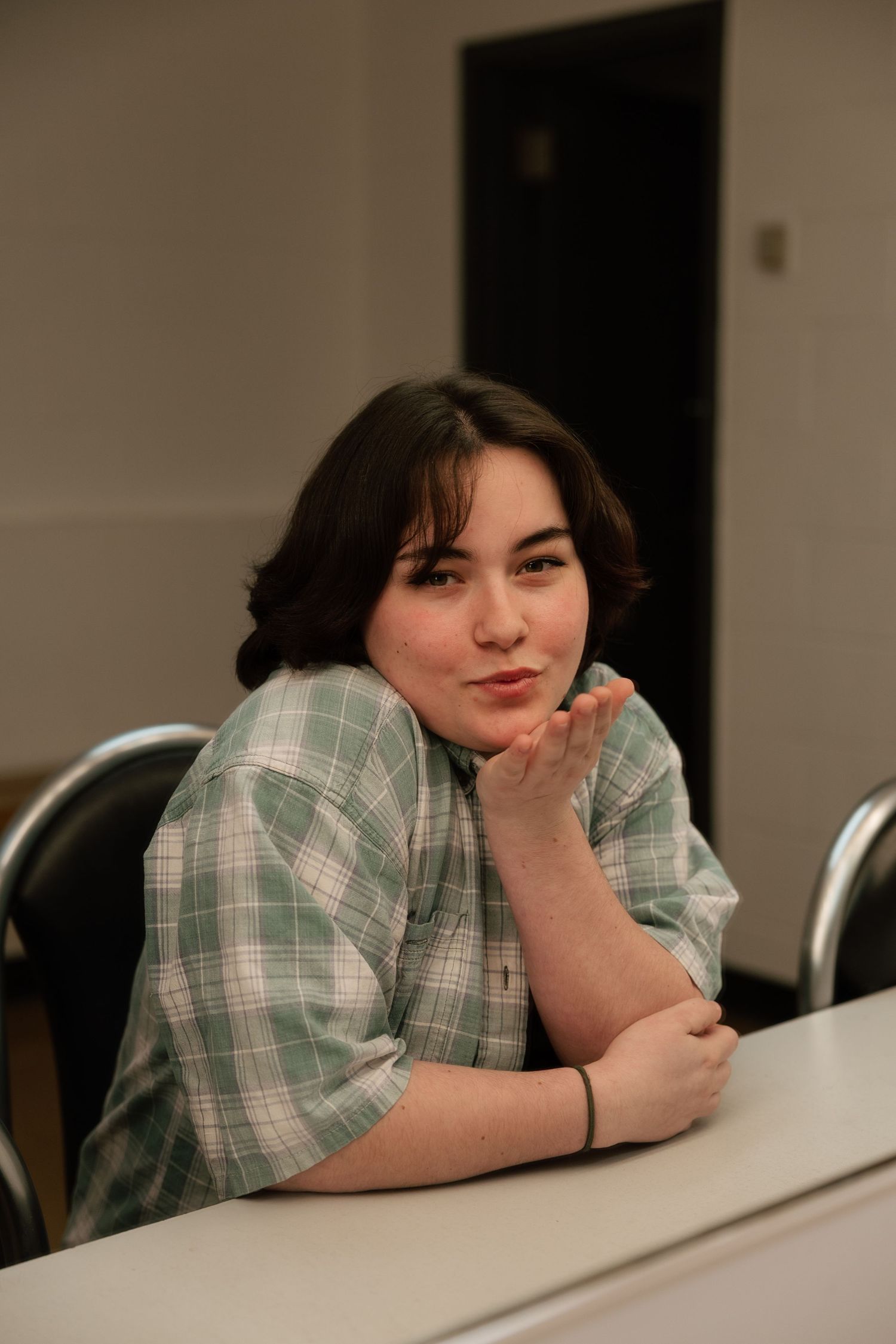 Young woman in green plaid shirt sitting at a table, resting her chin on her hand and smiling softly.