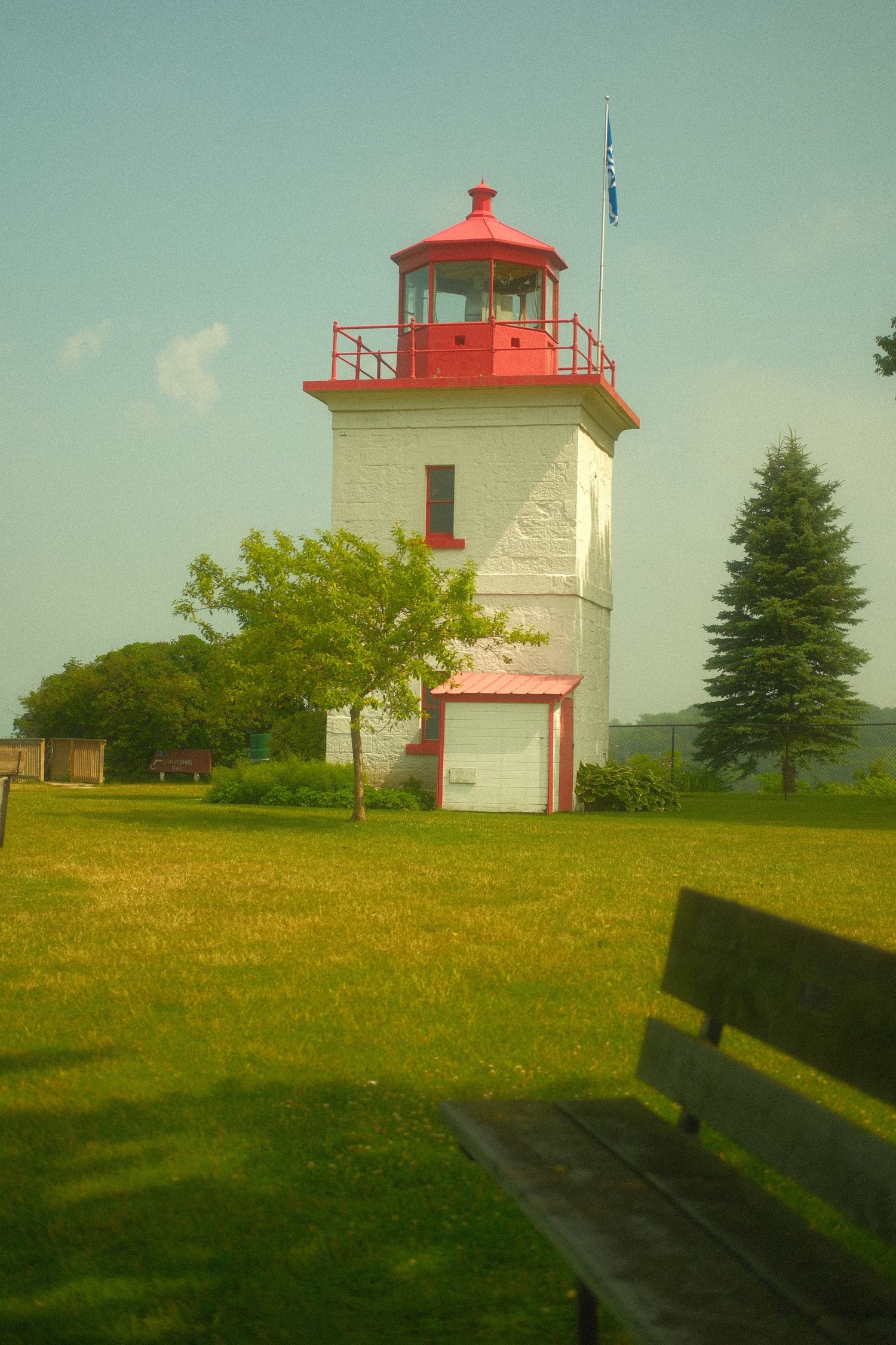 White and red lighthouse with a flag on a green lawn beside a wooden park bench.