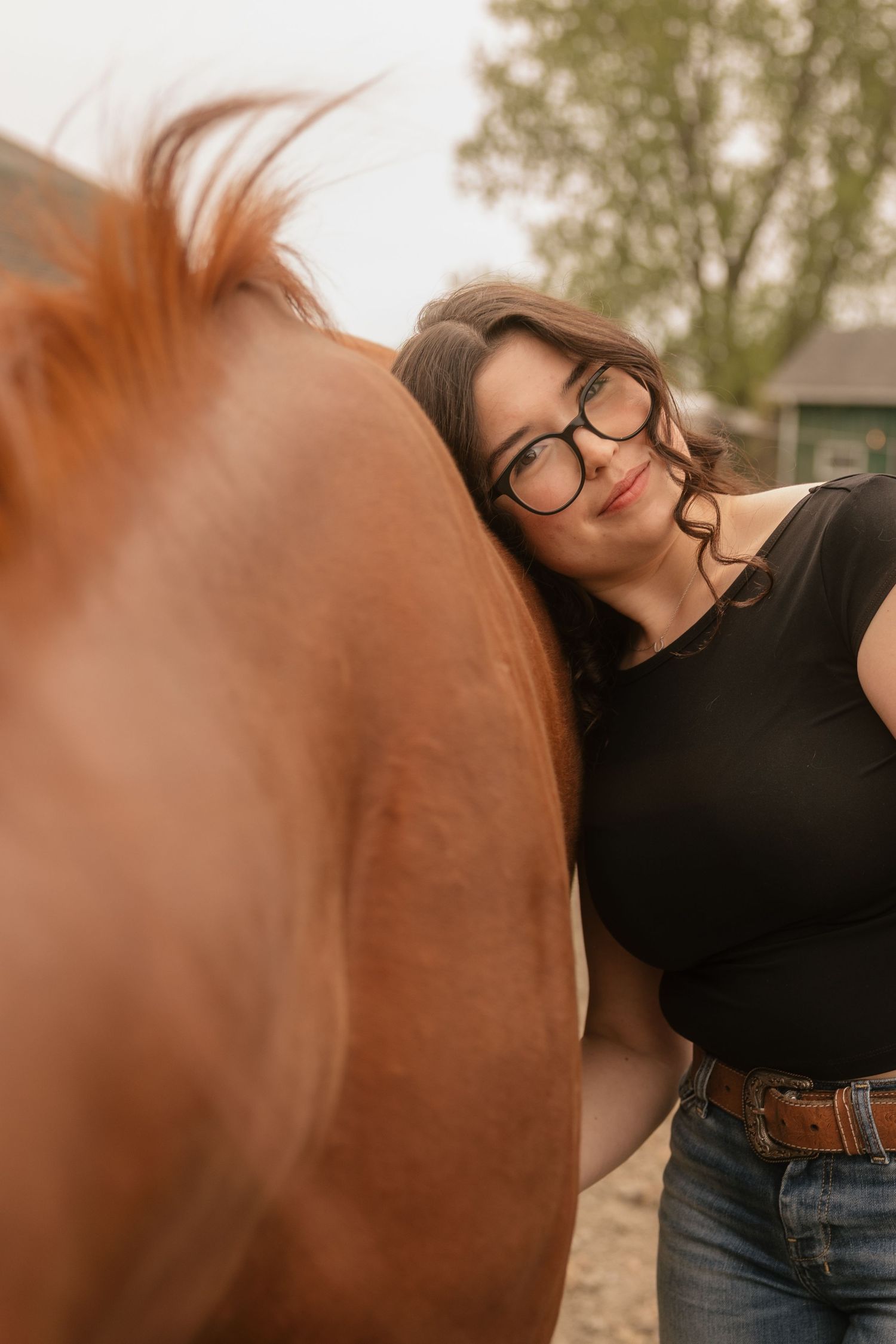 Young woman with glasses leaning affectionately against a chestnut horse outdoors near a barn.