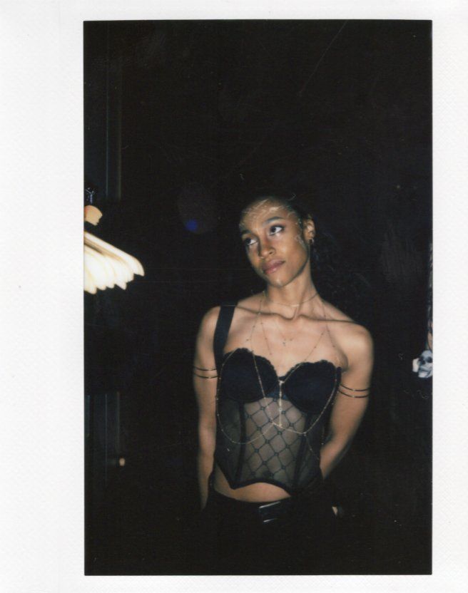 A young woman in a black mesh corset top poses in a dimly lit room in a Polaroid-style photo.