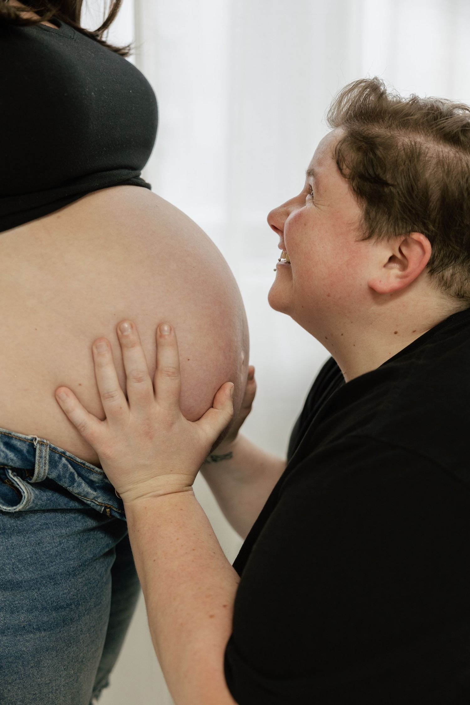 A smiling person tenderly touches a pregnant woman's bare baby bump during a maternity photo session.