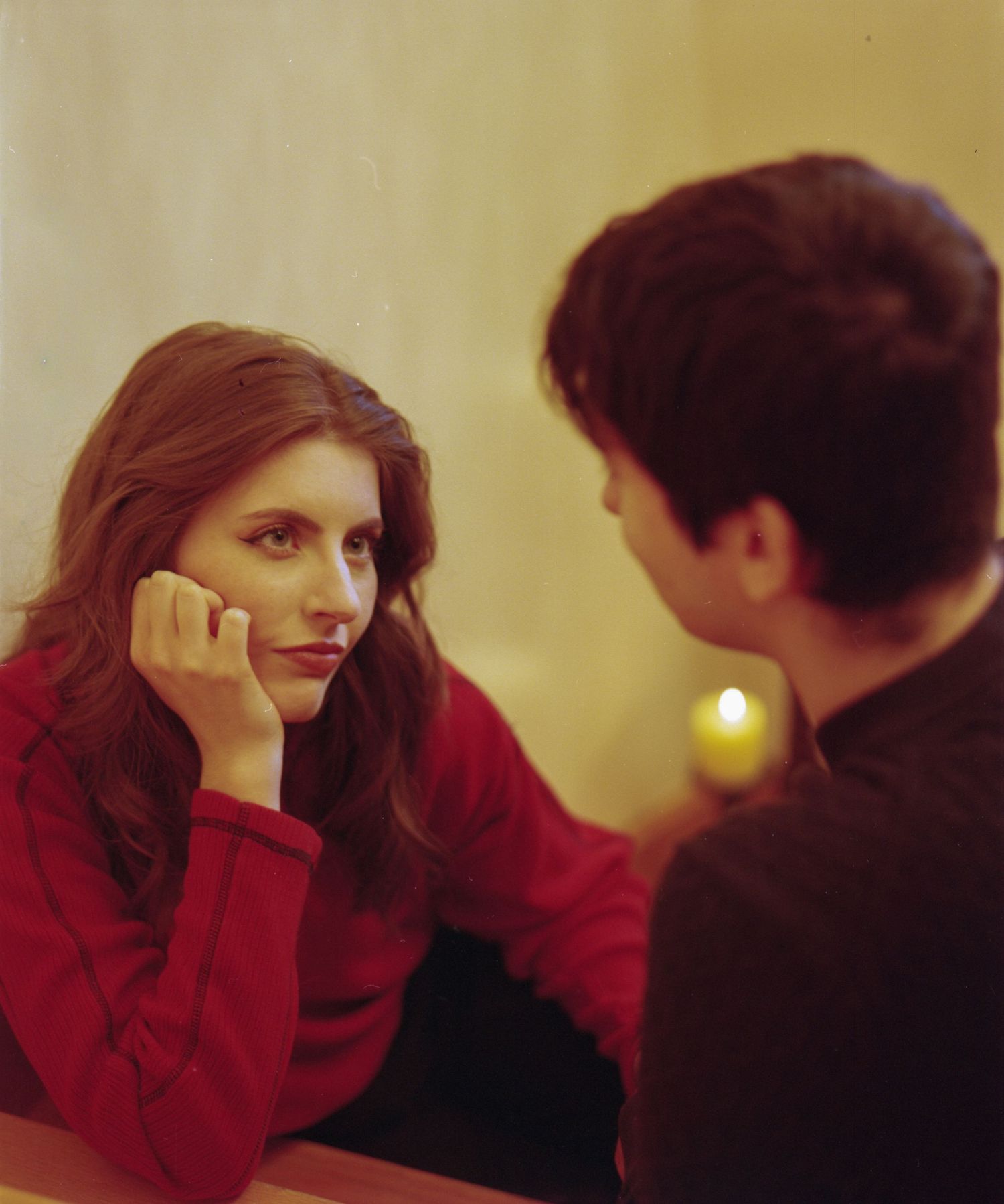 A woman in a red sweater gazes thoughtfully at a man by candlelight at a dinner table.