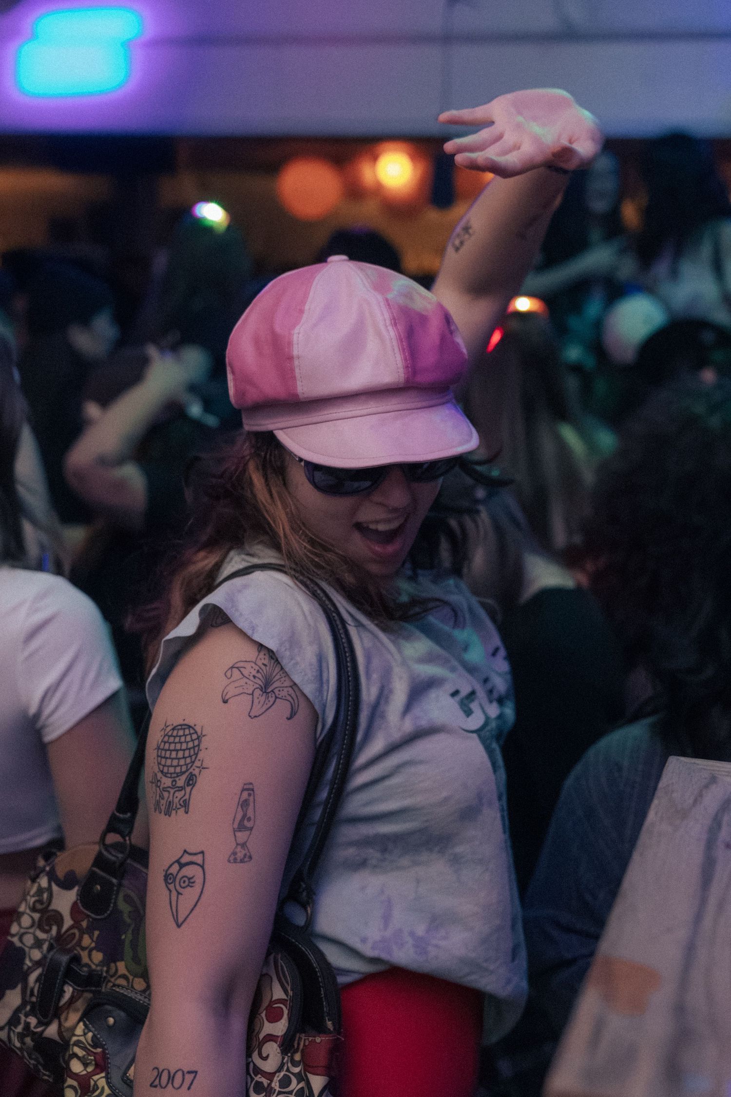 A woman in a pink cap with arm tattoos dances energetically at a crowded nightclub.