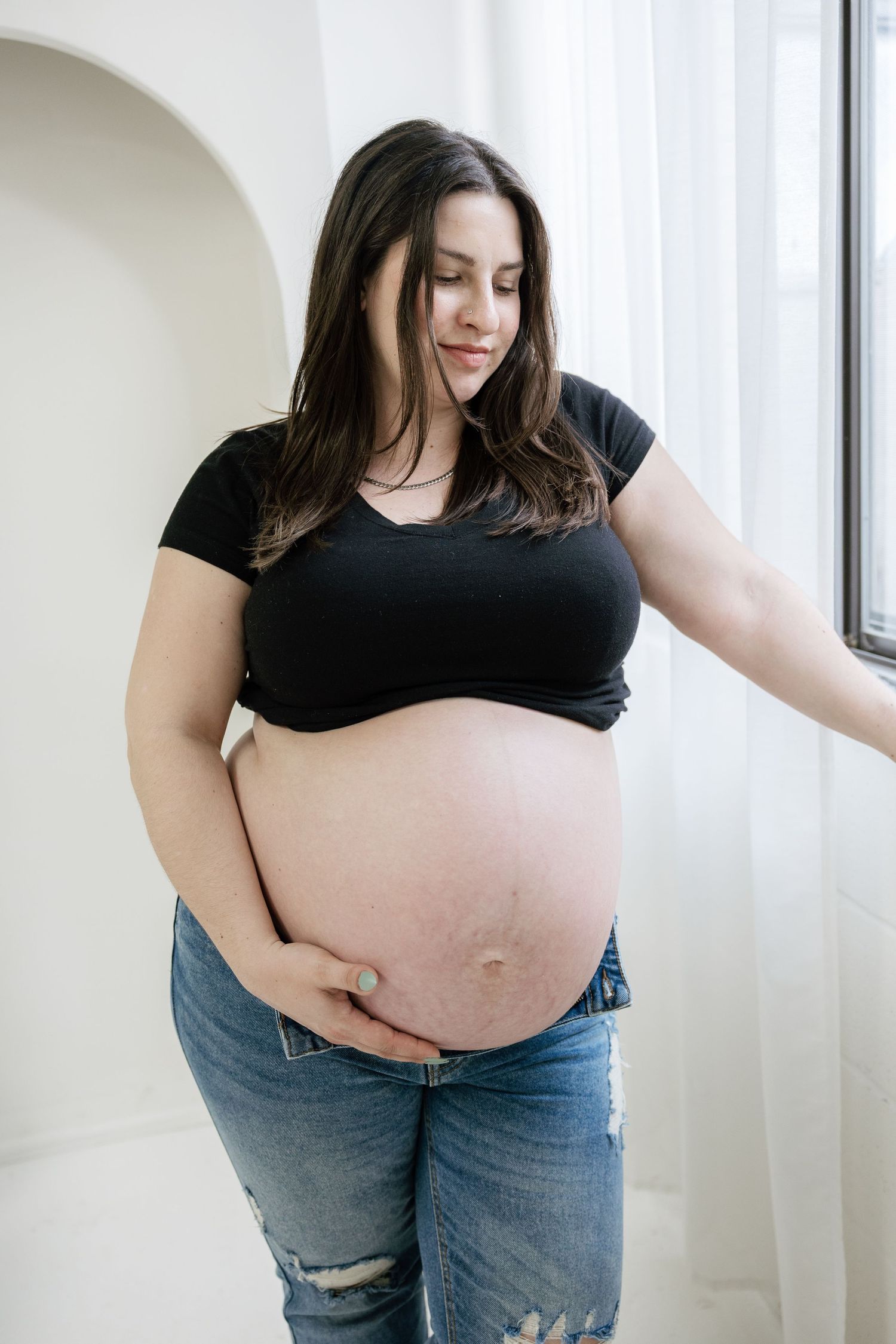 Pregnant woman in black crop top and jeans cradling her bare baby bump while standing near a bright window.
