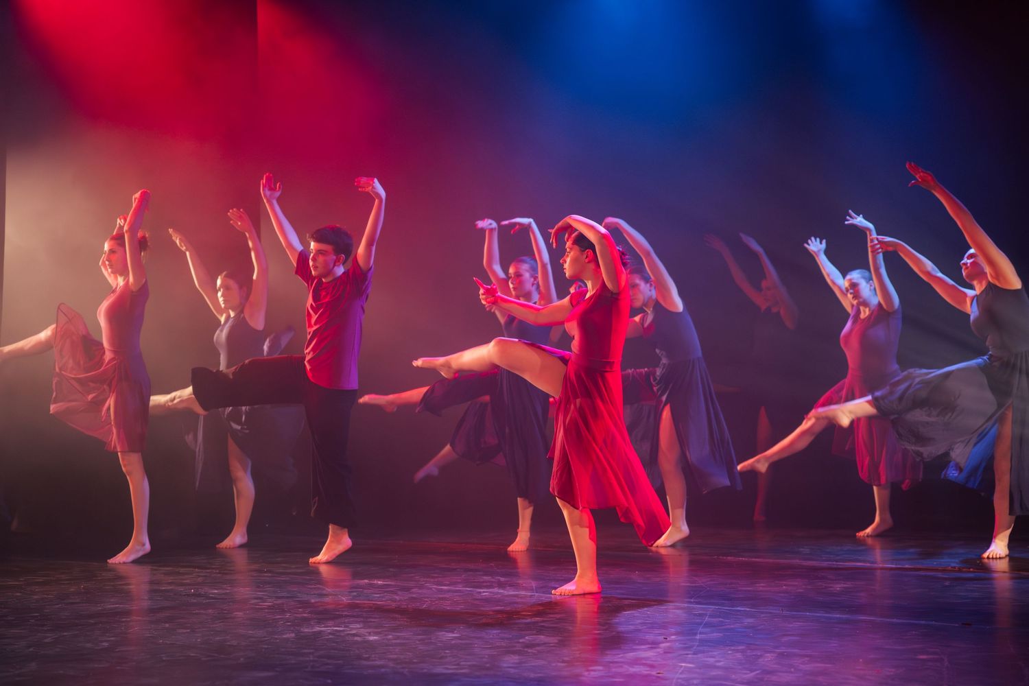 Contemporary ballet dancers performing arabesques in colorful costumes under red and blue stage lighting.