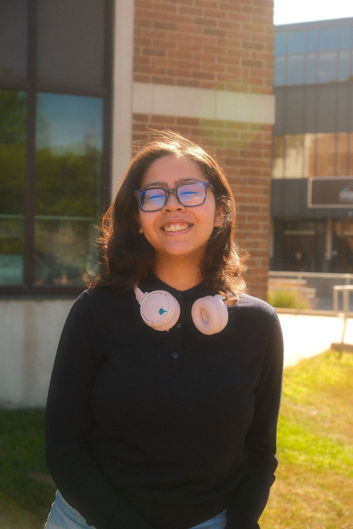Smiling young woman with glasses and pink headphones around neck, wearing black sweater outdoors near brick building.