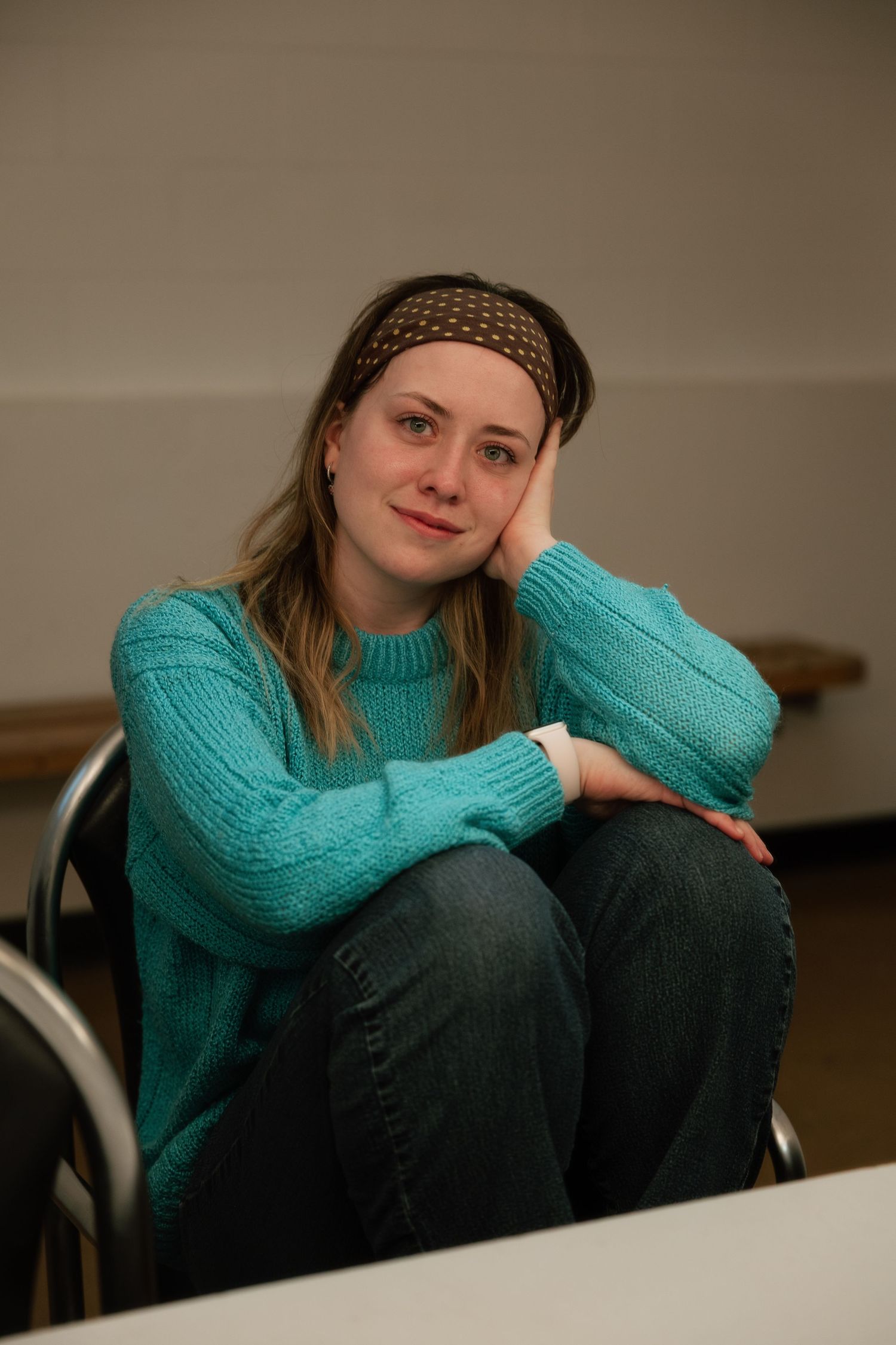 Young woman in teal sweater and headband sitting relaxed with hand on cheek in casual indoor setting.