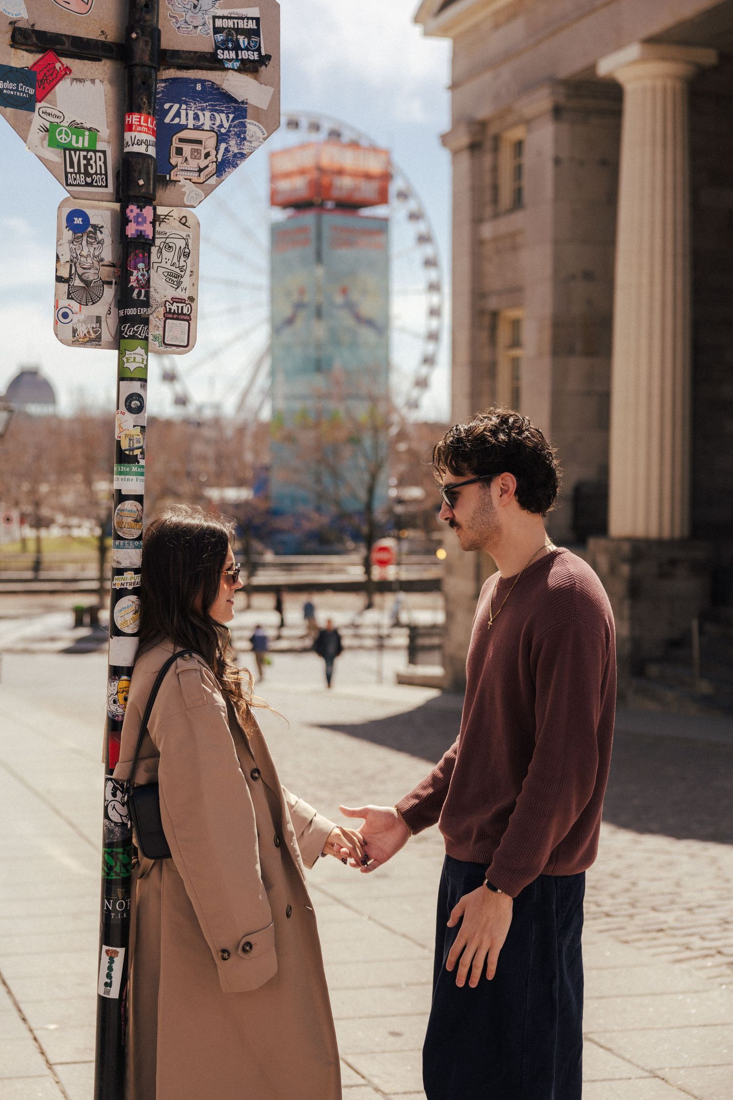 Couple holding hands near sticker-covered pole with Ferris wheel in background on sunny day.