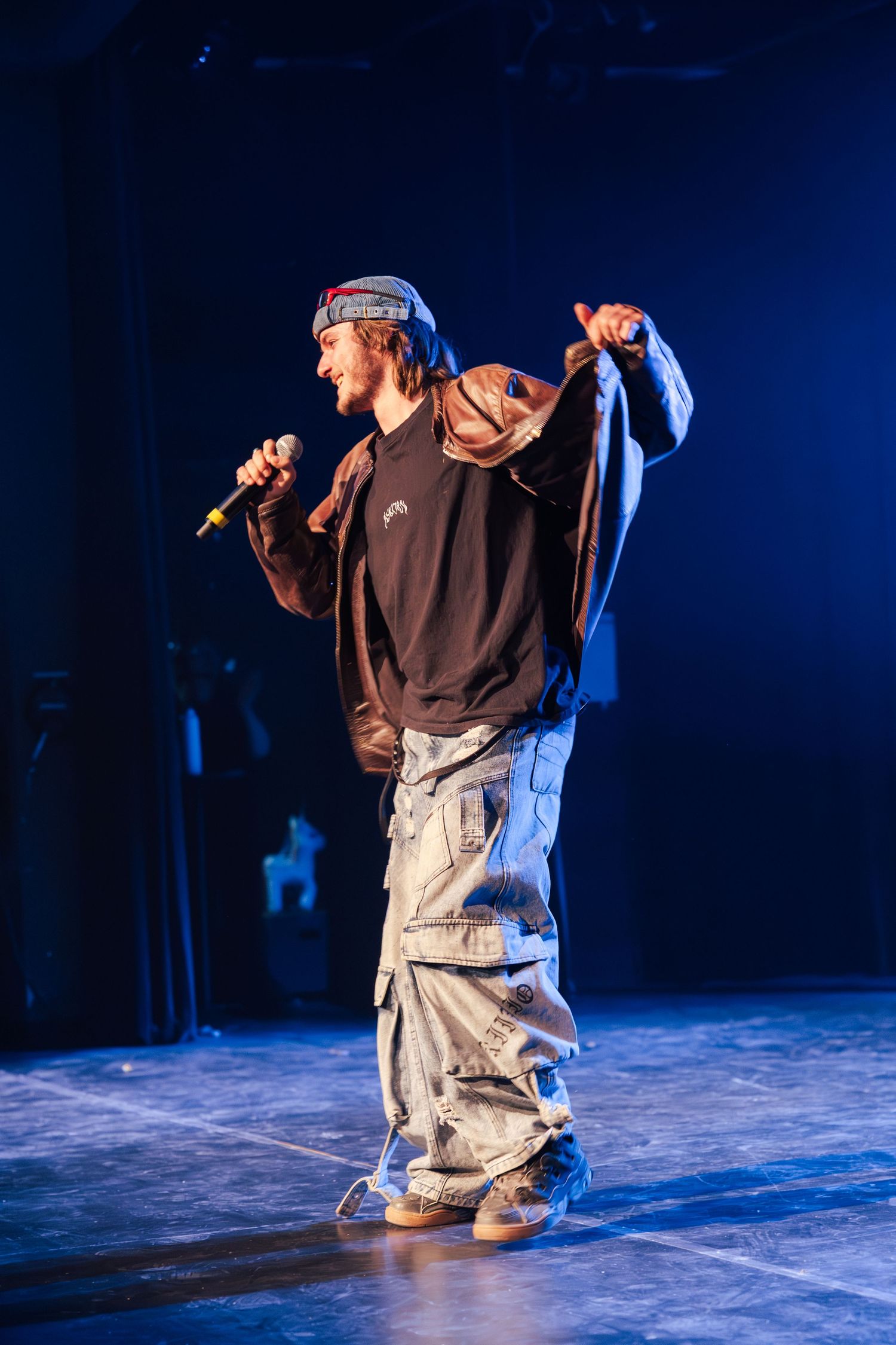 Male hip-hop artist performing on stage holding microphone wearing baggy cargo jeans and dark sweatshirt with blue lighting.