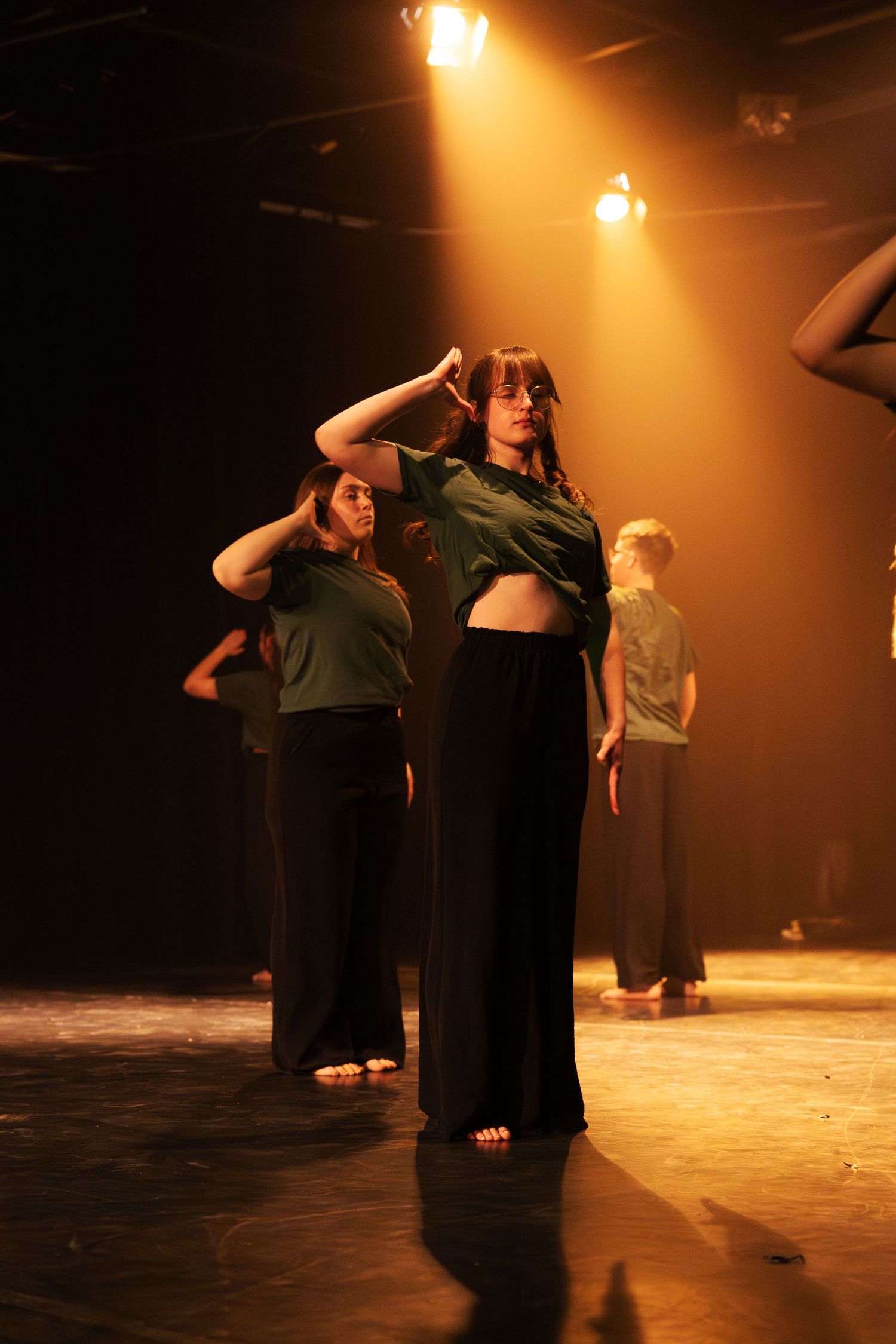 Dancers in green tops and black pants perform under warm amber stage lighting.