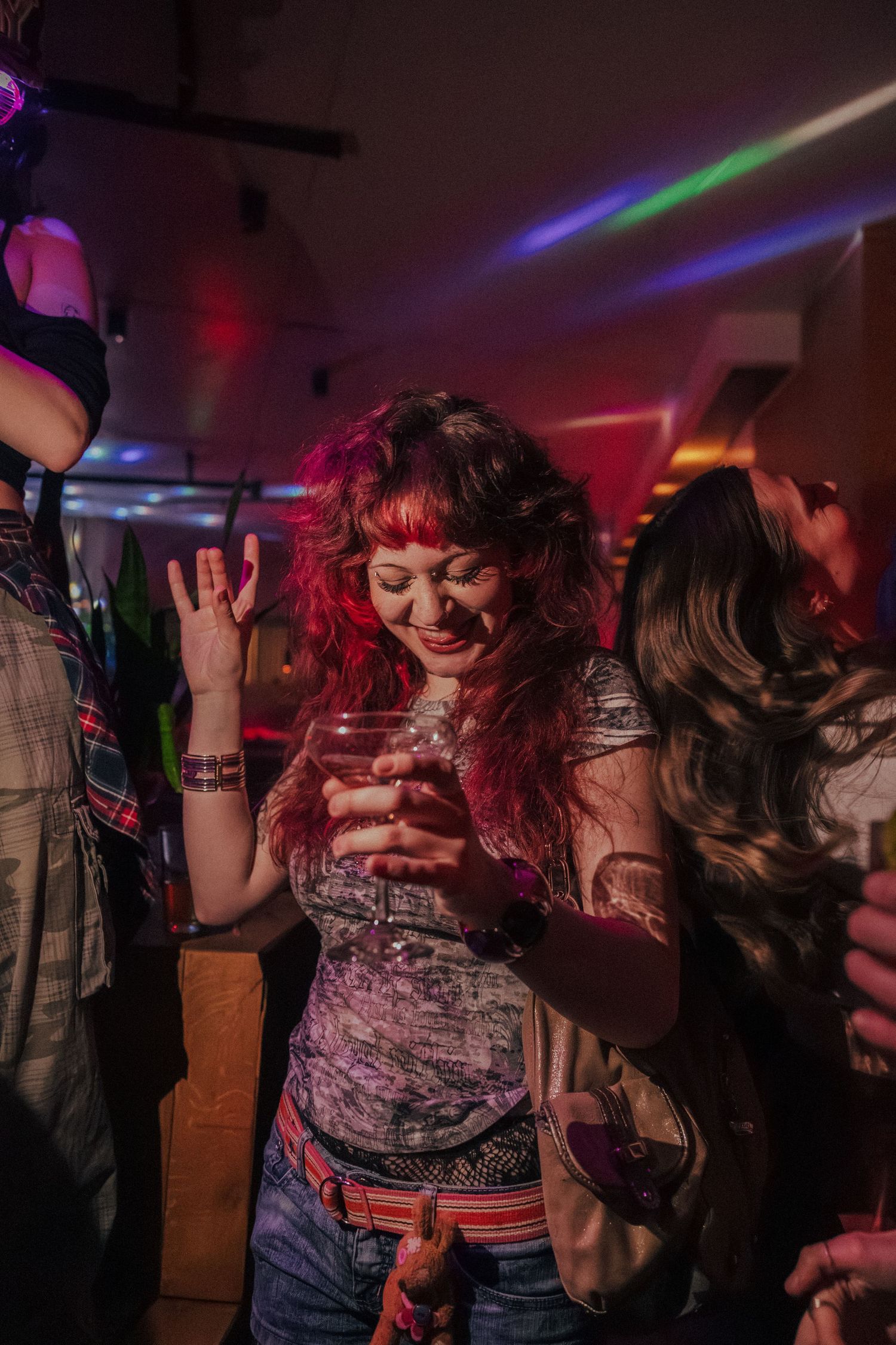 Woman with red curly hair dancing and holding a drink at a colorful nightclub.