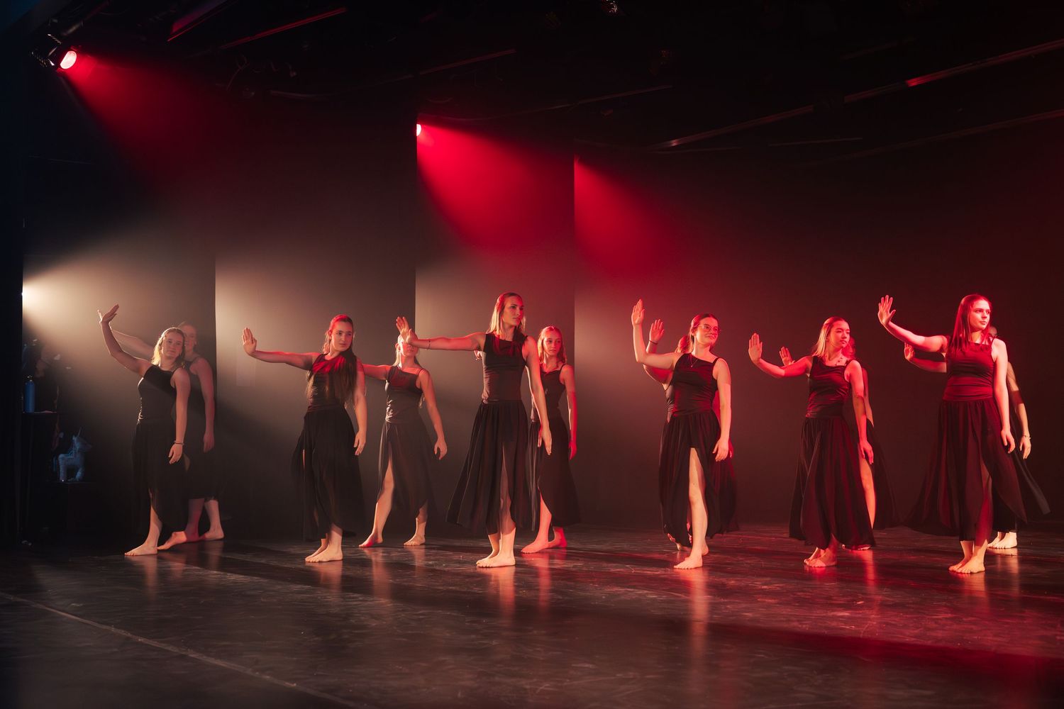 Line of dancers in black dresses performing synchronized routine with raised arms under dramatic red stage lighting.