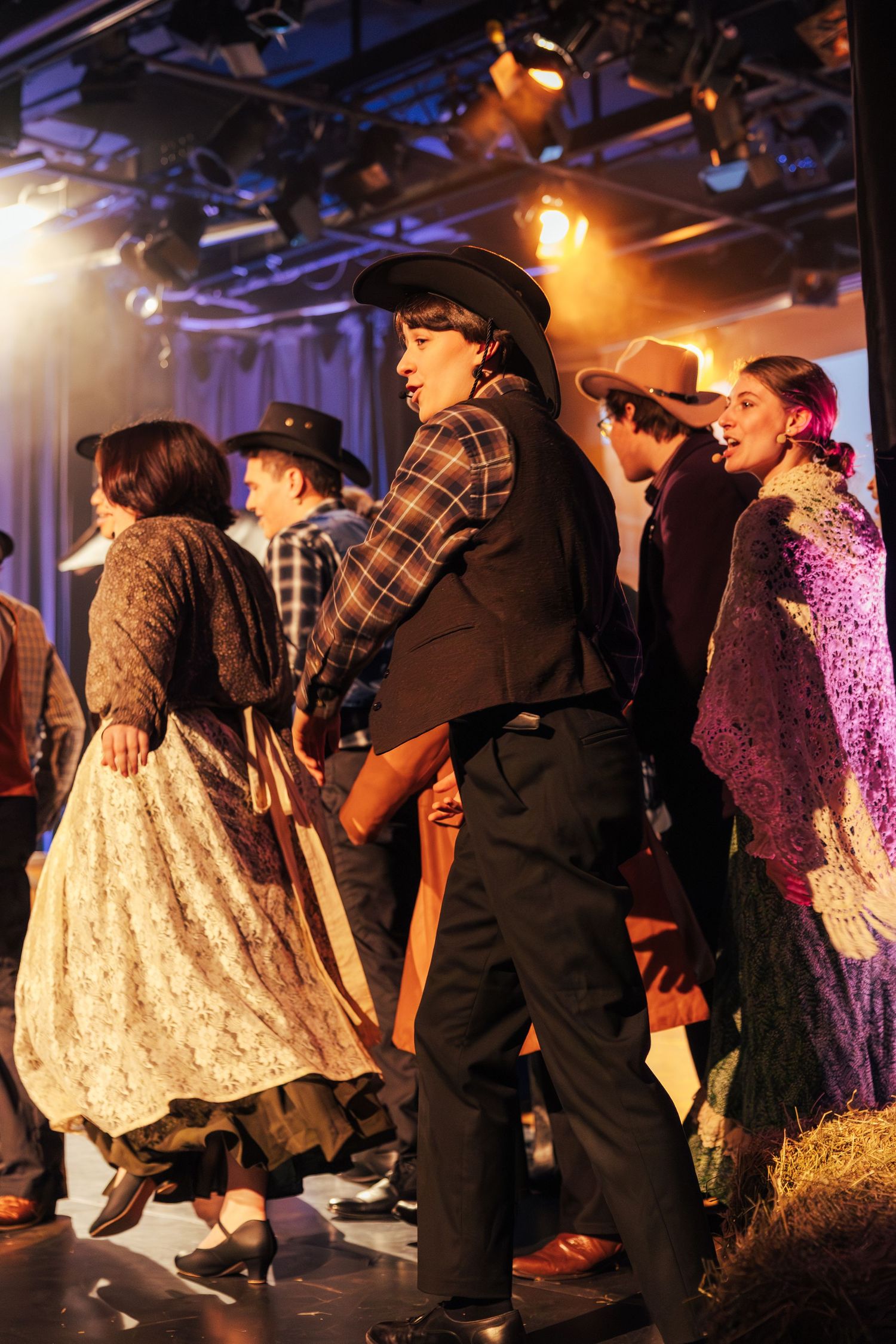 Performers in western cowboy costumes dance on stage with hay bales and warm golden stage lighting.