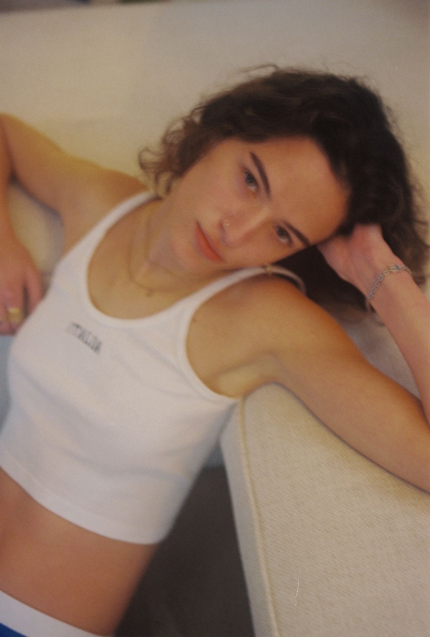 Young woman with curly dark hair resting on a sofa, wearing a white tank top, looking relaxed and casual.